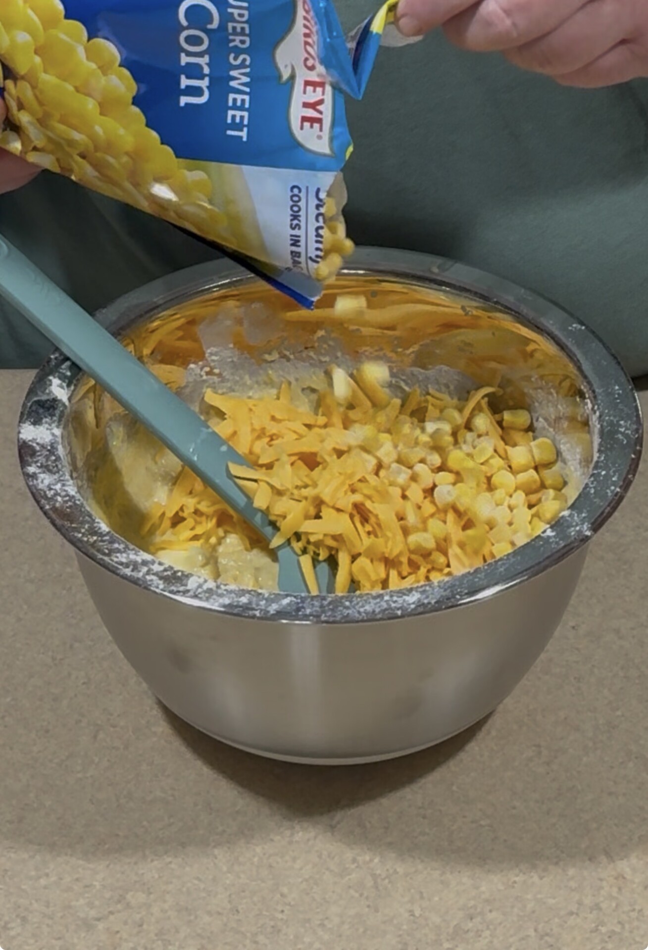 A person pours frozen corn from a bag into a mixing bowl filled with shredded cheddar cheese and dough batter, with a silicone spatula in the bowl.
