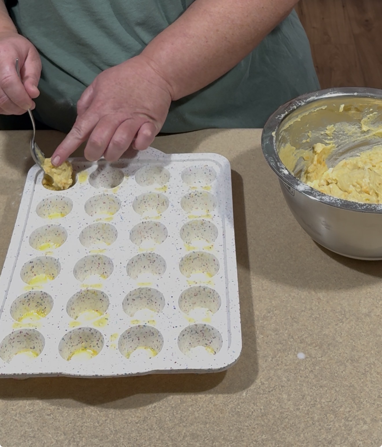 A person uses a spoon to scoop yellow batter from a metal bowl into a white, speckled silicone baking mold on a countertop.