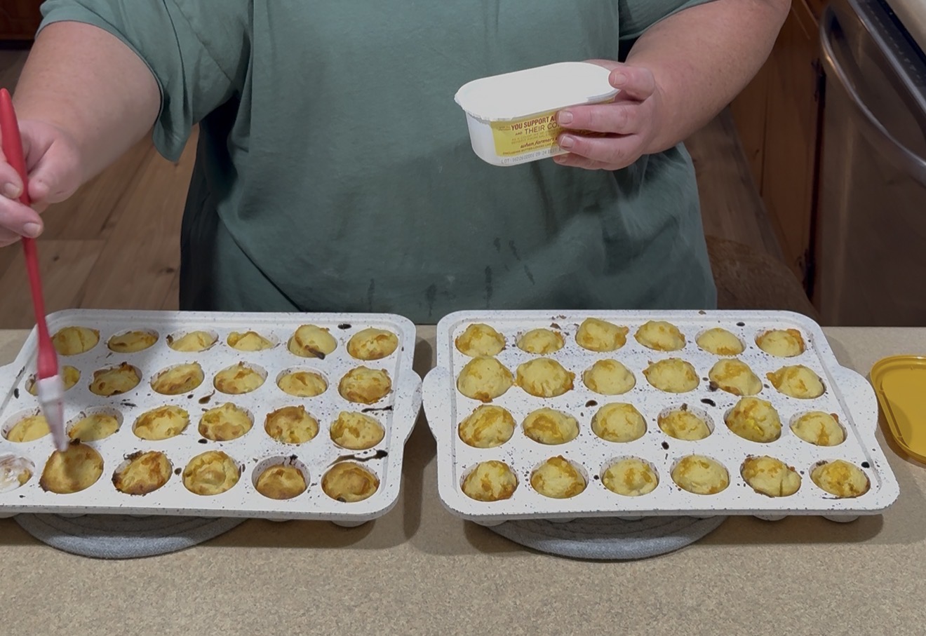 A person brushes melted butter onto small baked pastry shells in muffin trays, holding a butter container in one hand. The trays are on a kitchen counter.