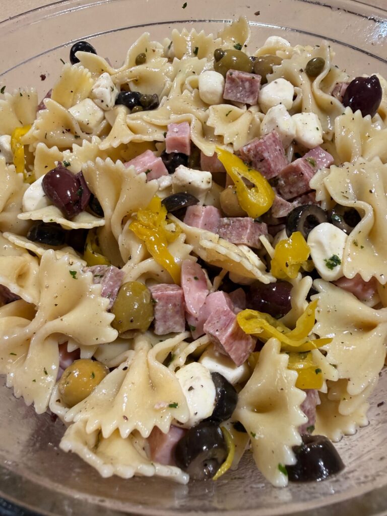 A bowl of Italian pasta salad with bowtie pasta, olives, mozzarella balls, cubed ham and salami, sliced pepperoncini, and Italian dressing, all mixed together.