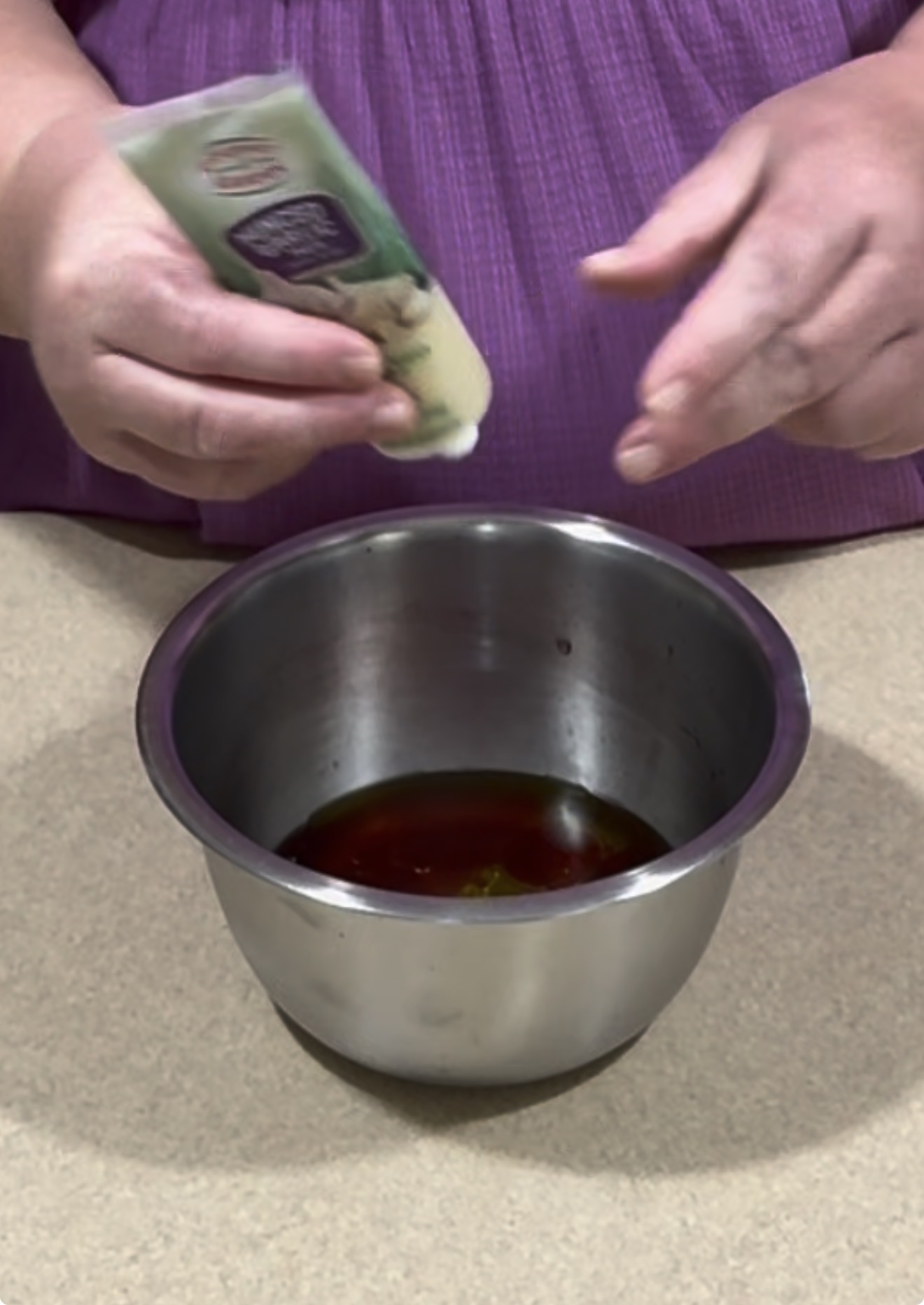 A person in a purple shirt squeezes a tube of seasoning or condiment into a metal bowl containing a dark liquid, on a beige countertop.