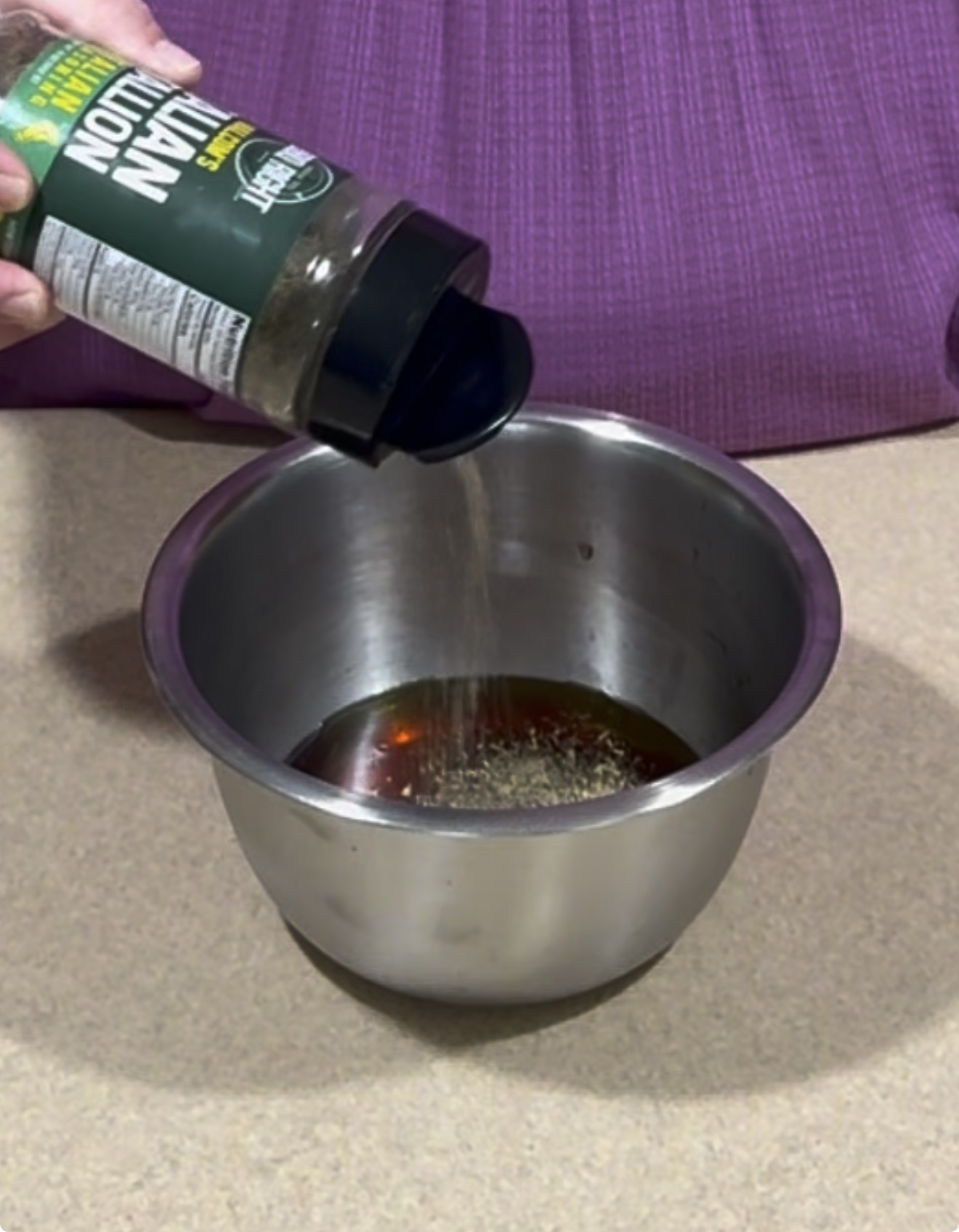 A hand sprinkles Italian seasoning from a container into a metal mixing bowl filled with liquid, on a countertop with a purple cloth in the background.