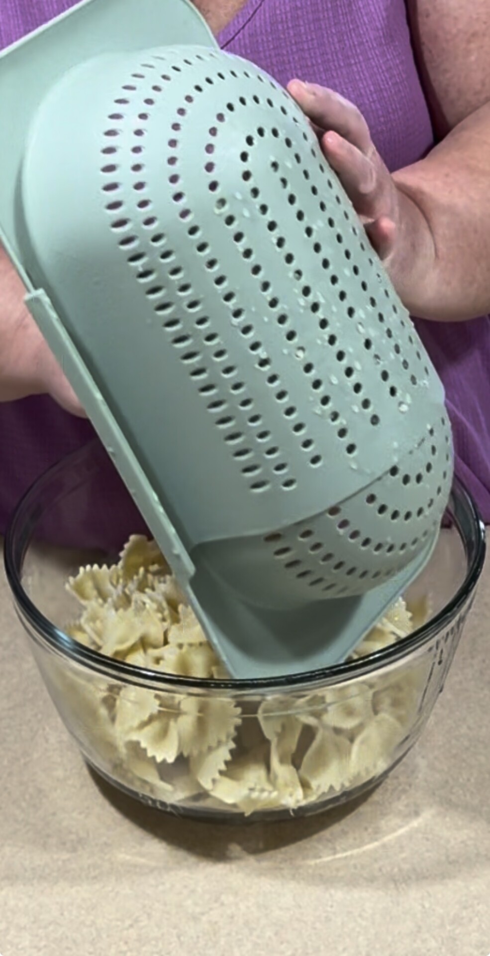 A person in a purple top pours cooked bowtie pasta from a green colander into a clear glass bowl.