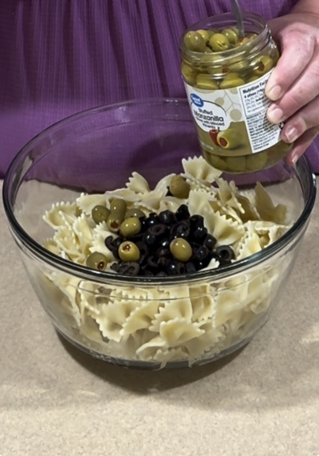 A person adds green olives from a jar into a large glass bowl filled with cooked bowtie pasta and black olives on a countertop.