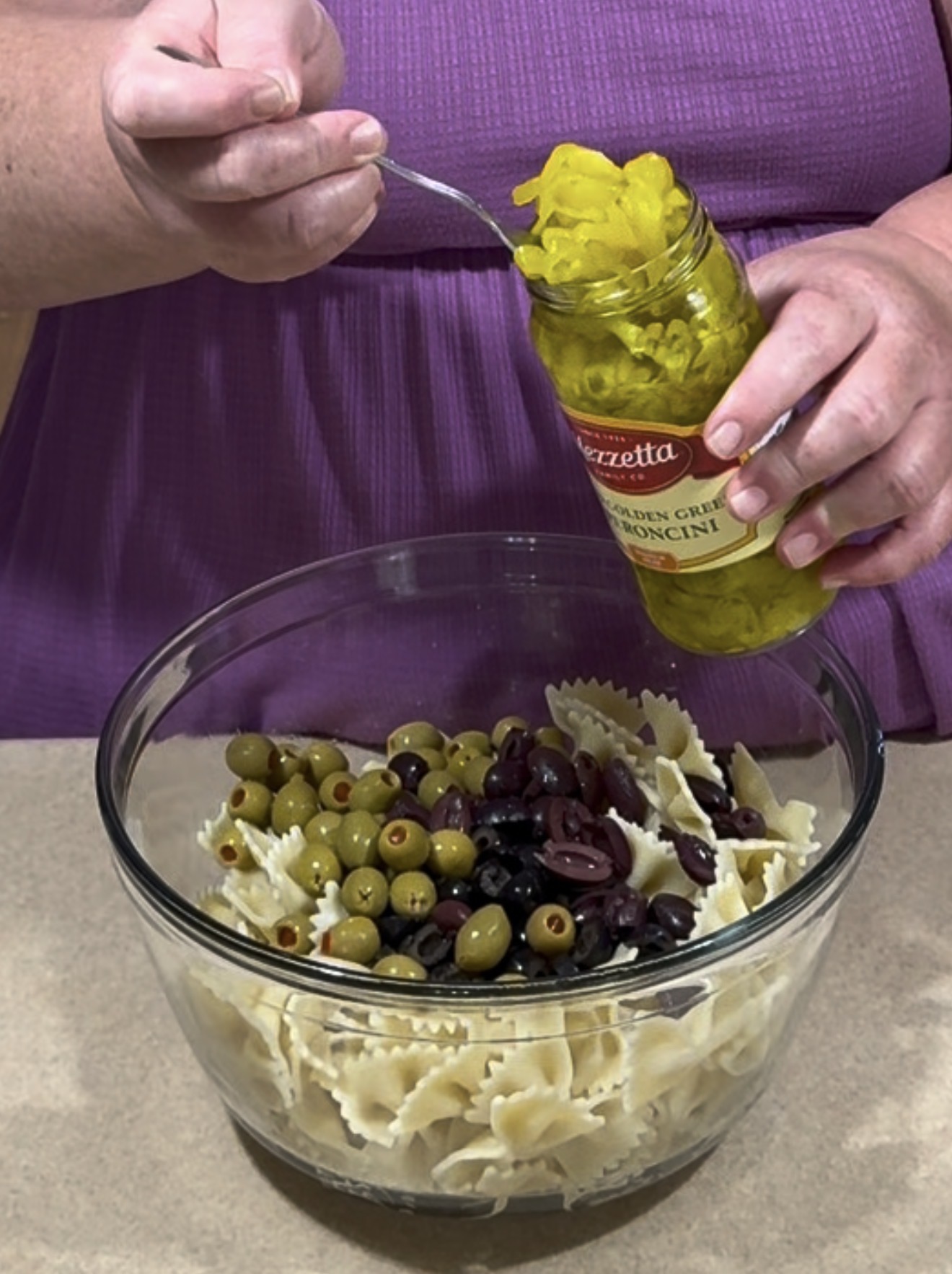 A person adds sliced pepperoncini from a jar to a glass bowl containing bowtie pasta and mixed olives. The person is wearing a purple shirt.
