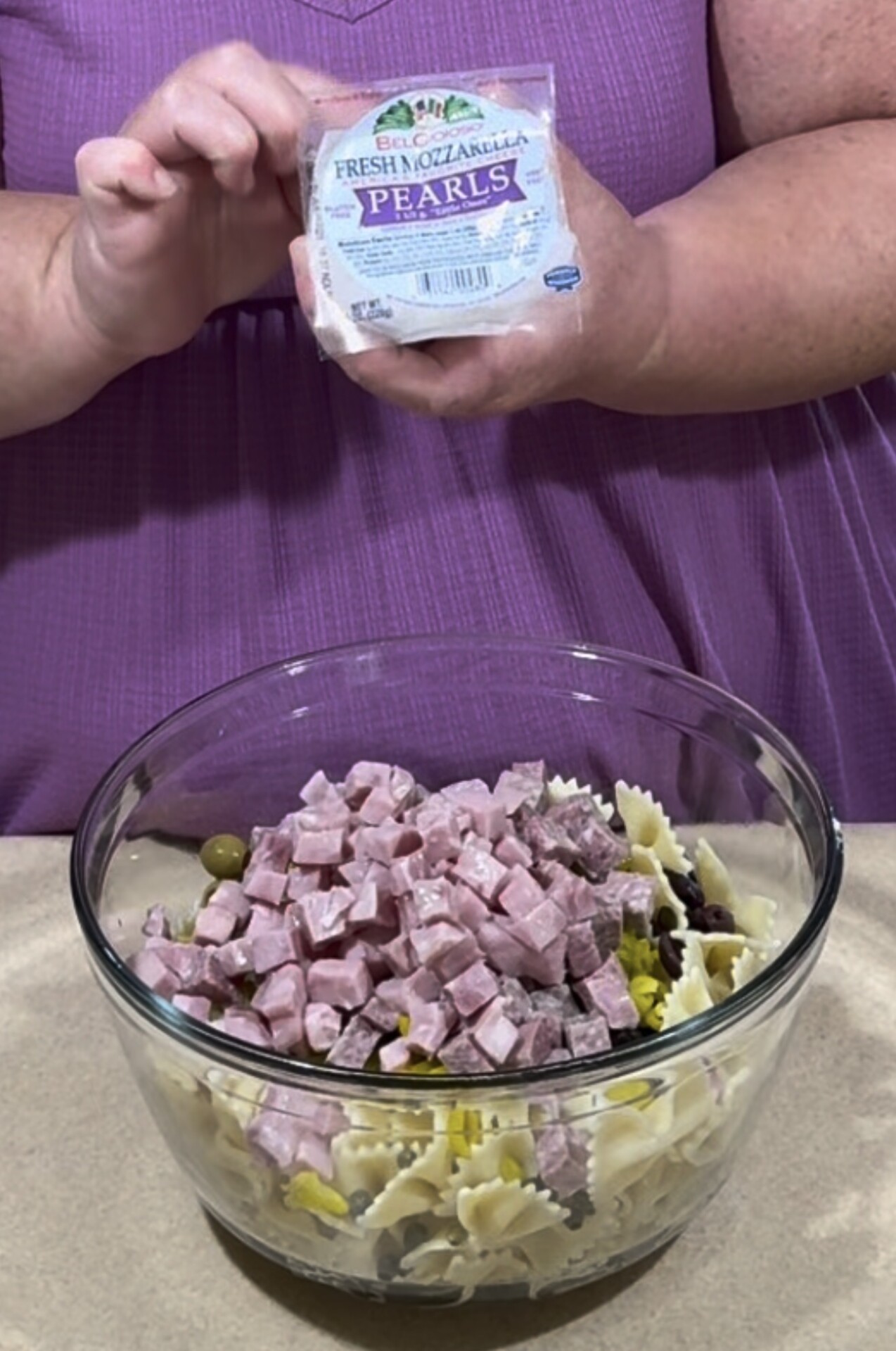 A person in a purple shirt holds a package of fresh mozzarella pearls above a glass bowl filled with bowtie pasta, cubed ham, olives, and other ingredients.