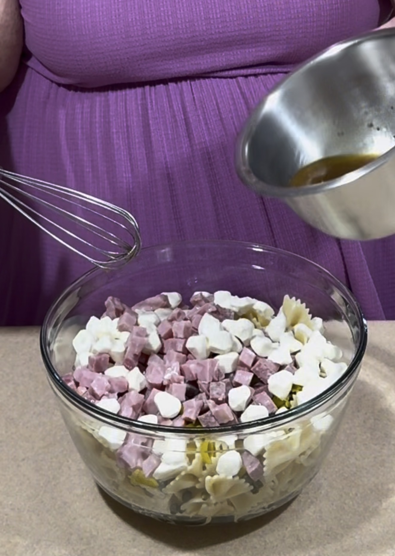 A person in a purple dress is preparing a pasta salad, holding a metal bowl with dressing over a glass bowl containing pasta, diced ham, and cheese cubes, with a whisk resting on the bowl.