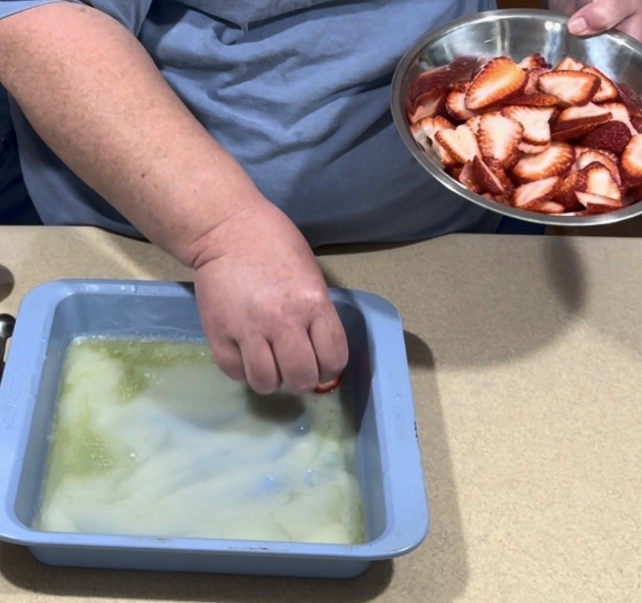 A person holds a metal bowl of sliced strawberries and places a slice into a blue dish filled with a white and greenish liquid on a beige countertop.