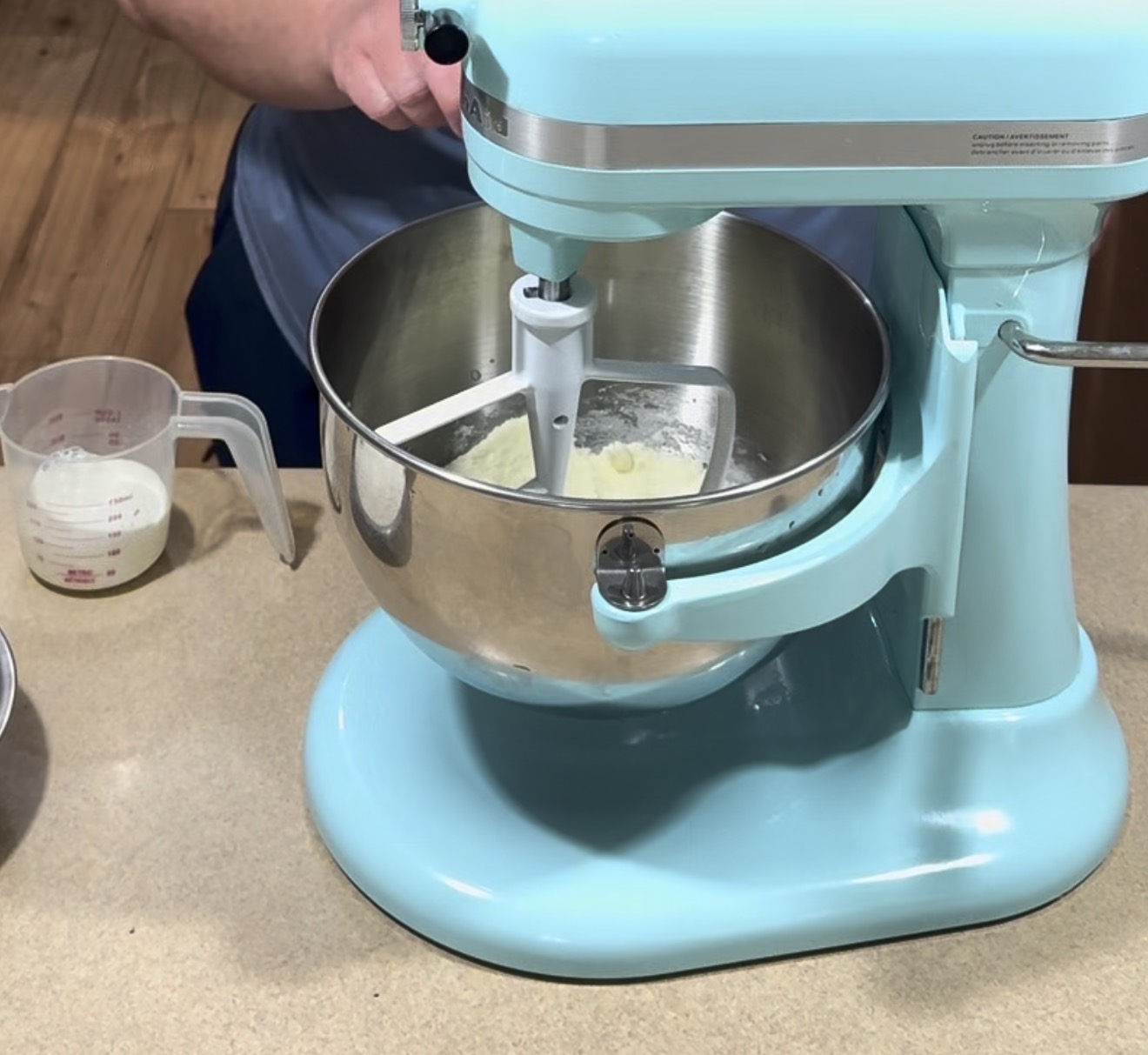 A person uses a light blue stand mixer to mix ingredients in a metal bowl. A measuring cup with liquid is on the countertop nearby. The scene appears to be in a kitchen.