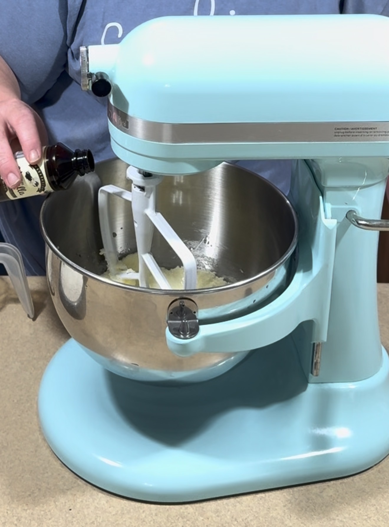 A person pours vanilla extract into a light blue stand mixer containing ingredients, preparing to mix them in a stainless steel bowl on a kitchen counter.
