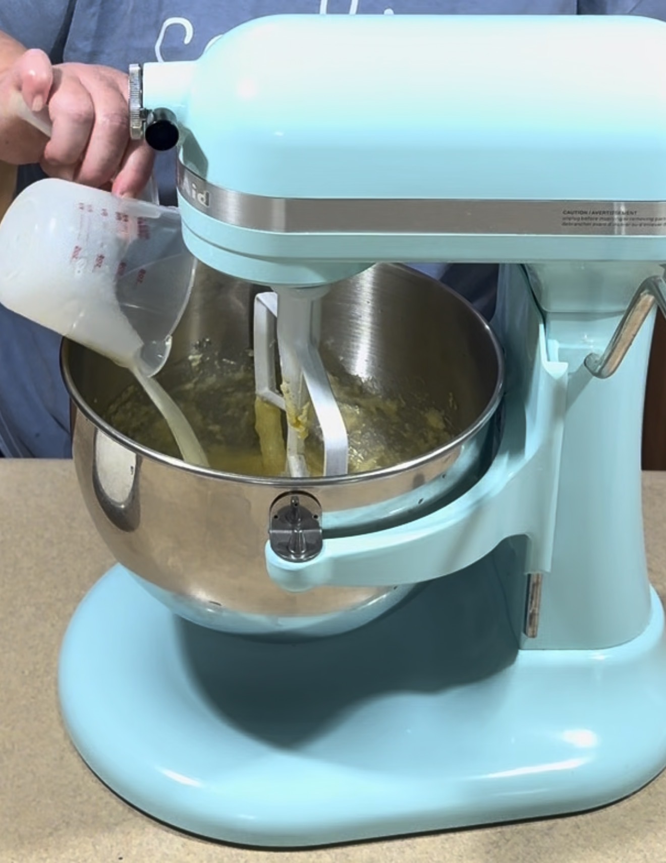 A person pours liquid from a measuring cup into a blue stand mixer that is mixing yellow batter in a metal bowl.