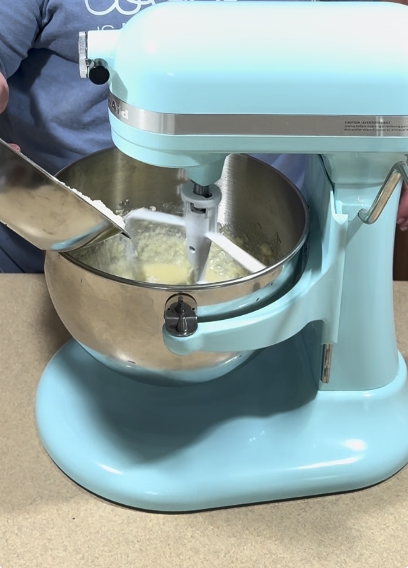 A person uses a spatula to scrape ingredients into a pale blue stand mixer with a metal bowl on a kitchen counter. The mixer is running and mixing a batter.
