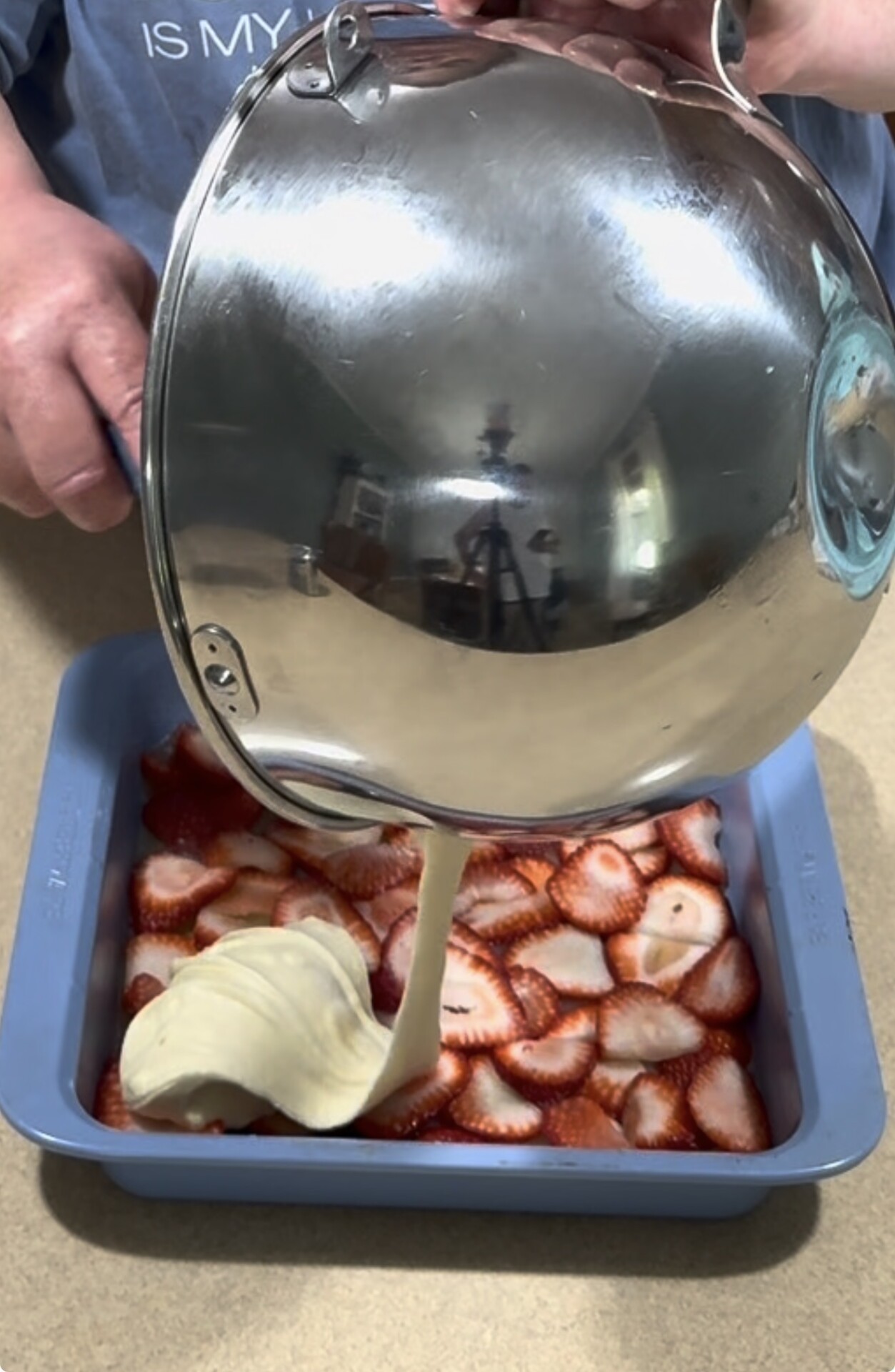 A person pours creamy batter from a metal mixing bowl over a layer of sliced strawberries in a blue square baking dish on a countertop.