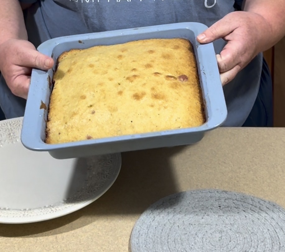 A person holds a square baking pan containing a freshly baked golden-brown cake, ready to be served. The pan is above a counter with a plate and a round textured mat nearby.