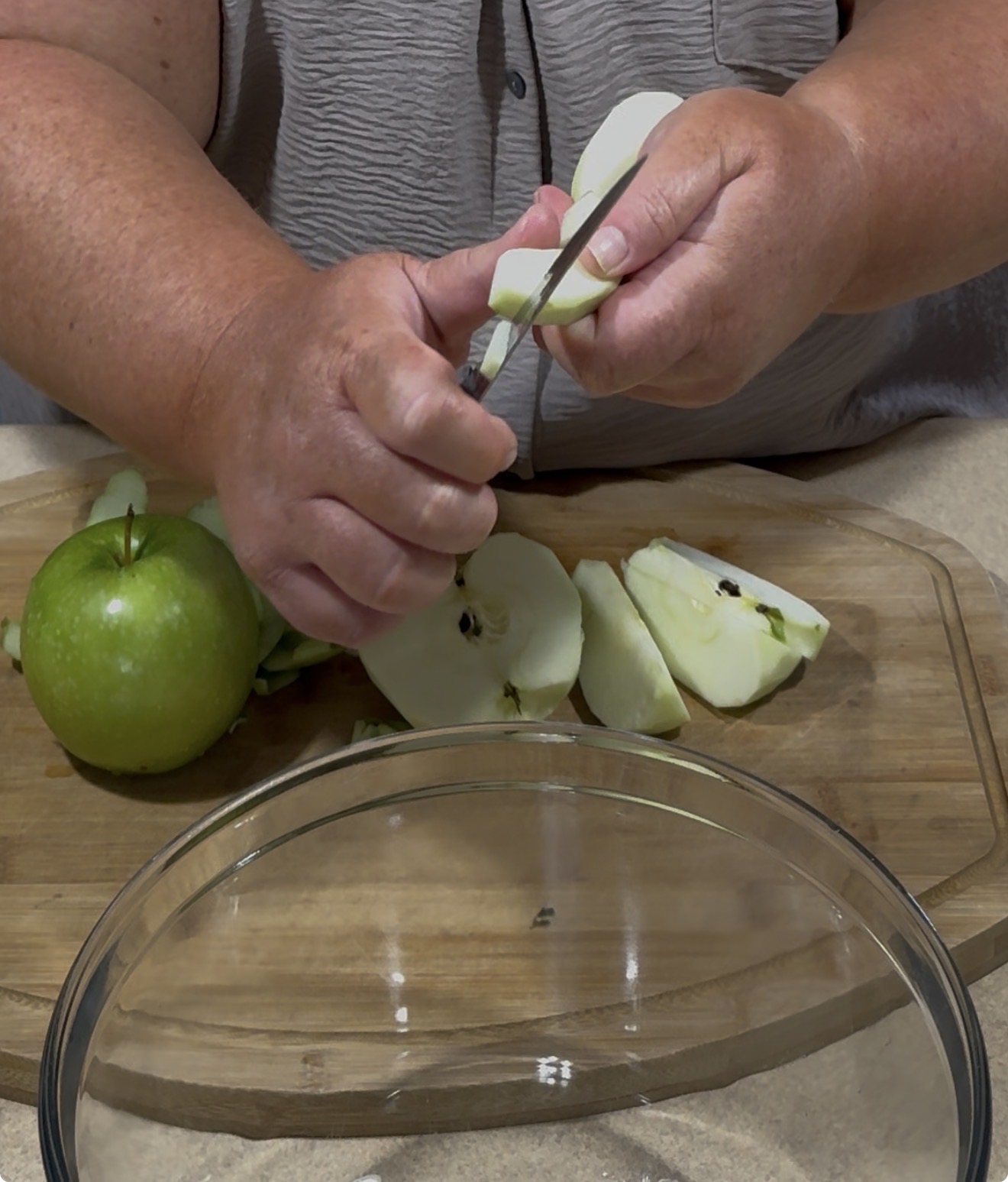 A person uses a knife to peel and slice a green apple on a wooden cutting board. More apple slices and a glass bowl are in the foreground. The person is wearing a light-colored shirt.