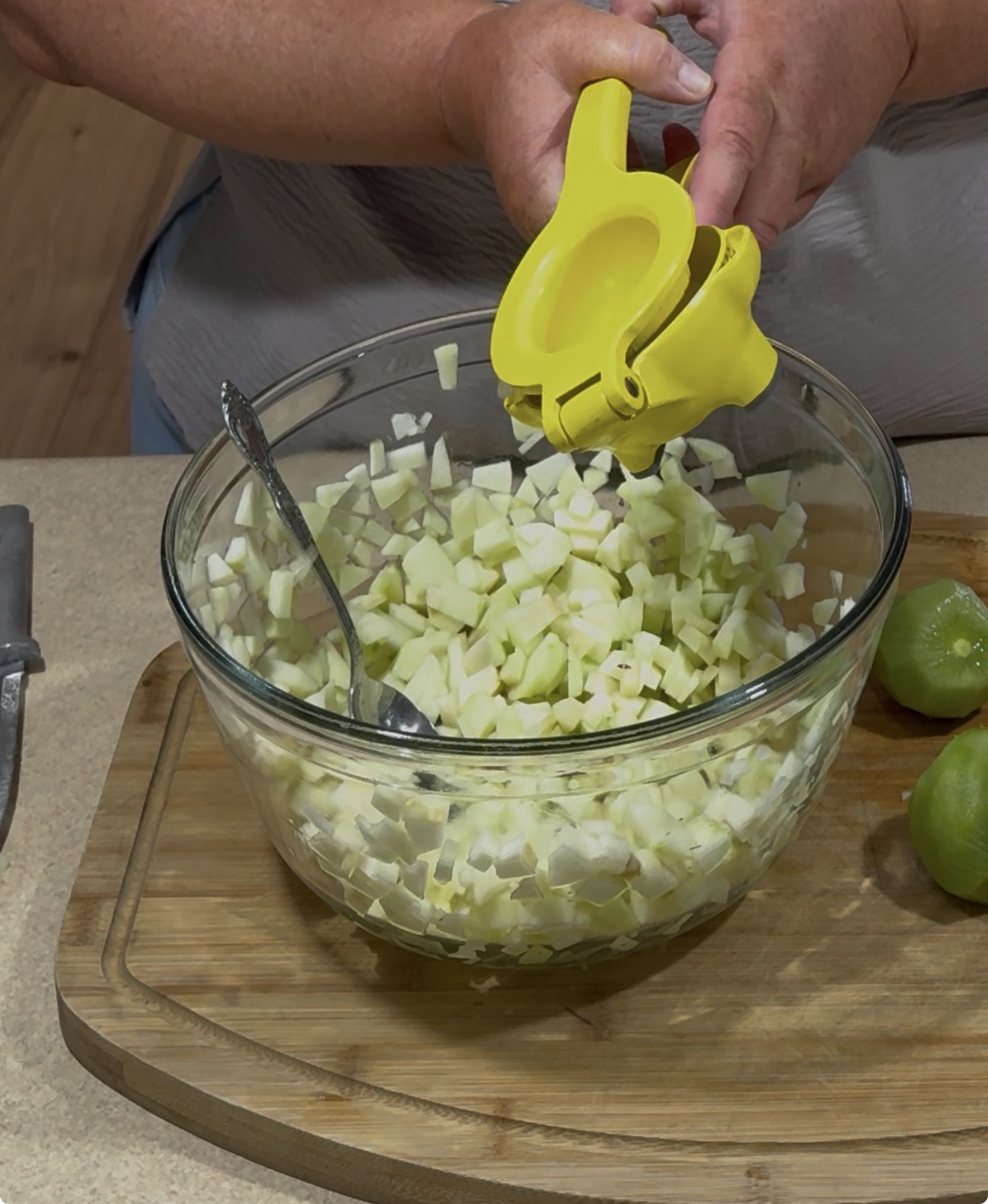 A person holds a yellow citrus squeezer over a glass bowl filled with chopped green apples on a wooden cutting board, with a spoon in the bowl and halved limes nearby.