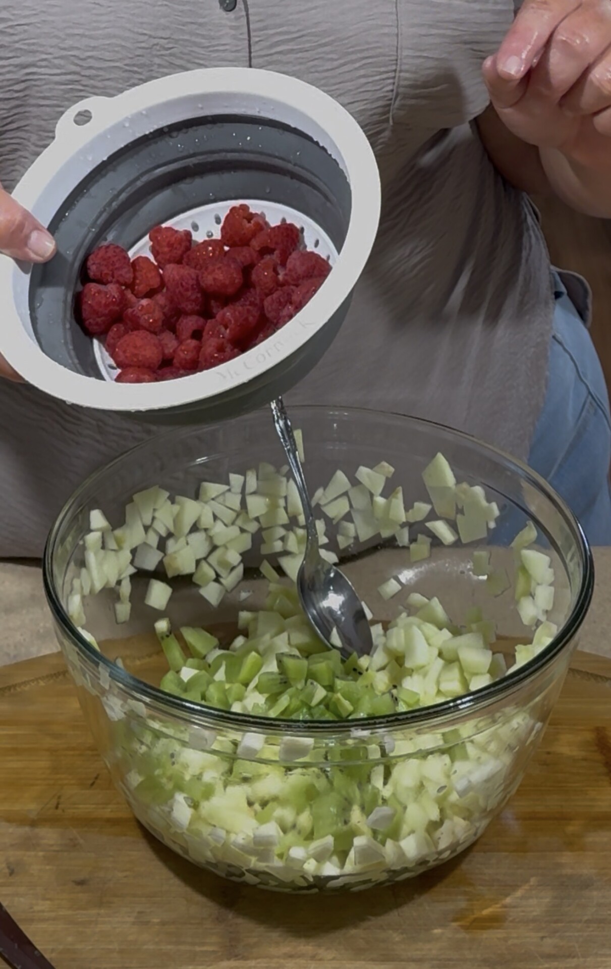 A person holds a strainer with raspberries above a glass bowl containing diced green apples and celery, with a spoon resting inside the bowl.