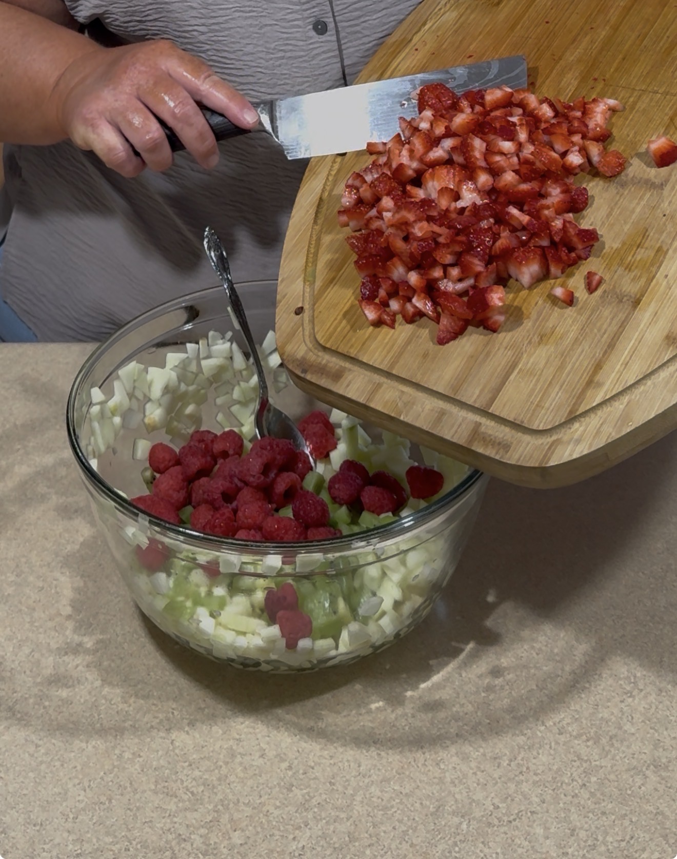 A person adds chopped strawberries from a cutting board into a glass bowl containing diced apples, green grapes, and raspberries on a kitchen counter.