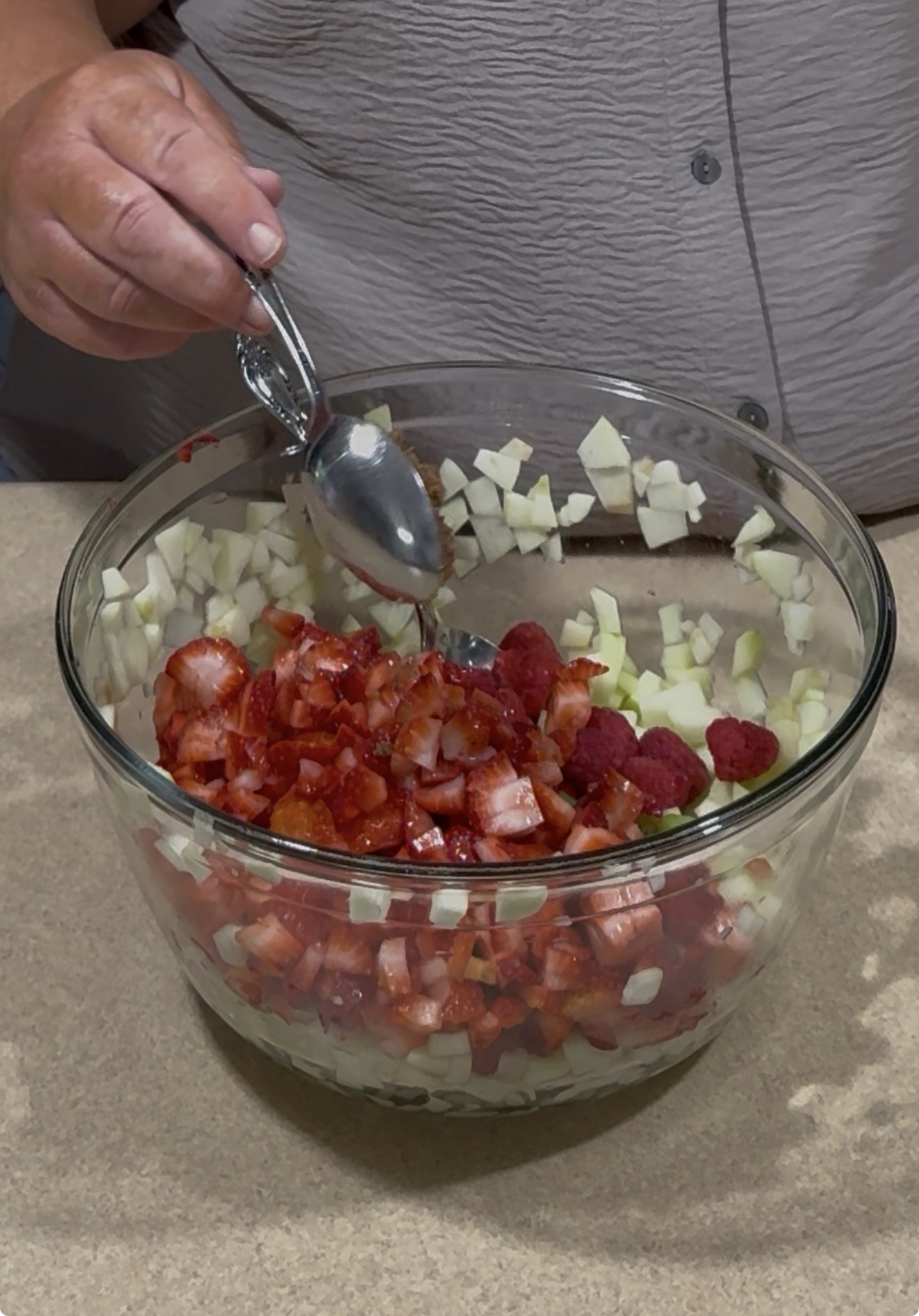 A person holding two spoons prepares to mix chopped apples, strawberries, and raspberries in a large glass bowl on a countertop.