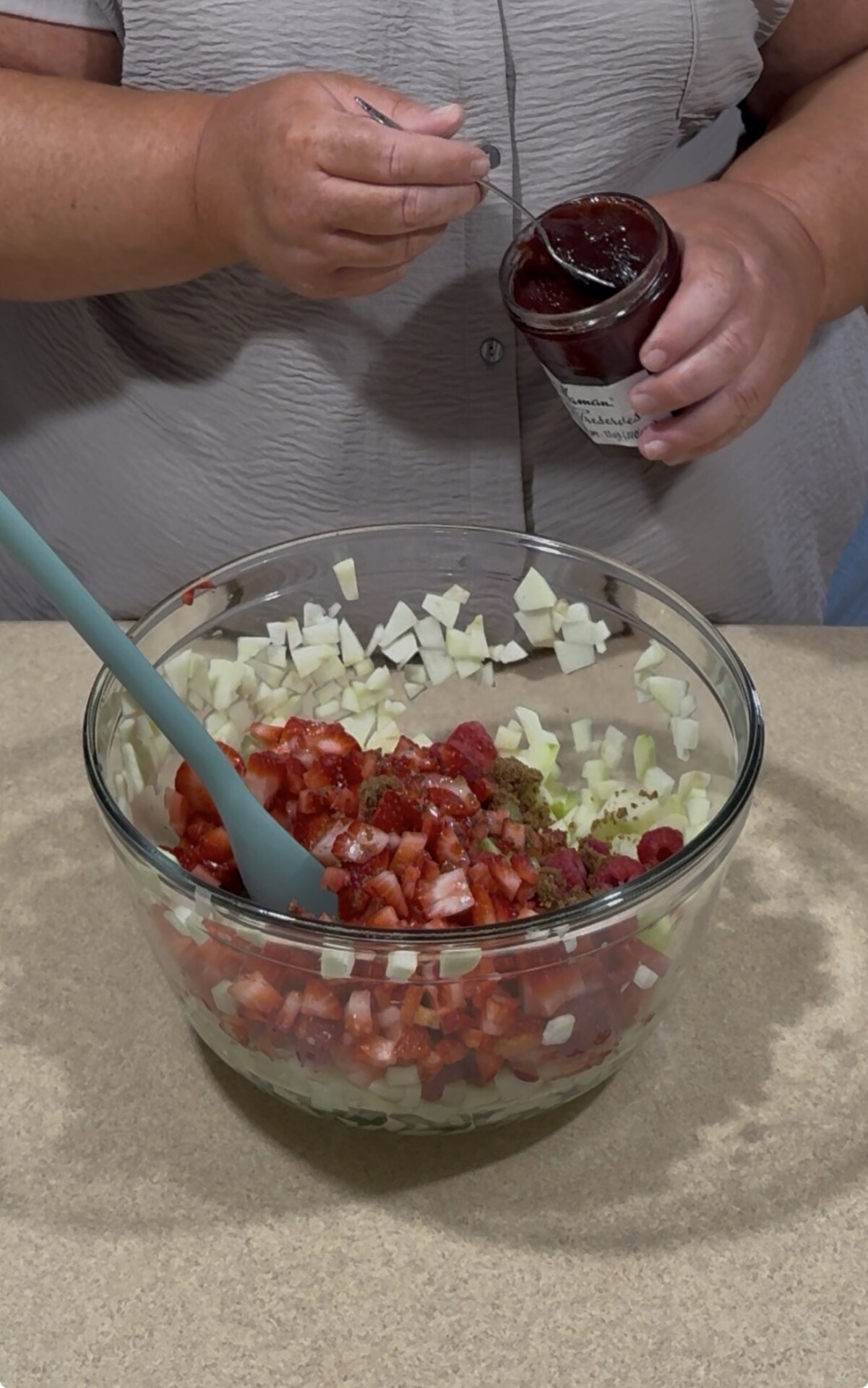 A person adds strawberry jam from a jar into a glass bowl filled with chopped onions, strawberries, and other ingredients on a kitchen counter. A blue spatula is in the bowl, and the person wears a gray shirt.