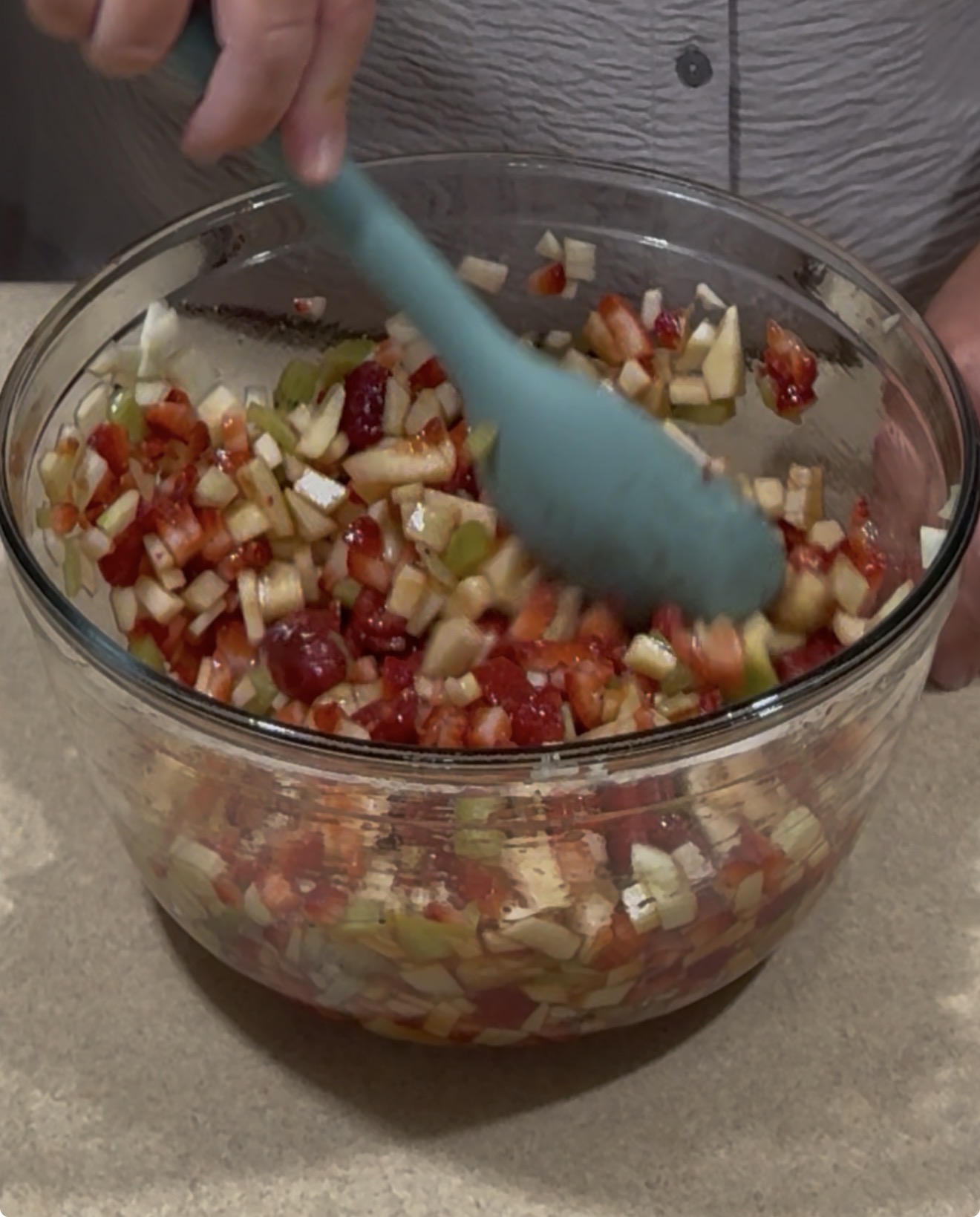 A person stirring a colorful fruit salad with a blue spatula in a clear glass bowl on a kitchen counter. The salad contains chopped apples, strawberries, and other assorted fruits.