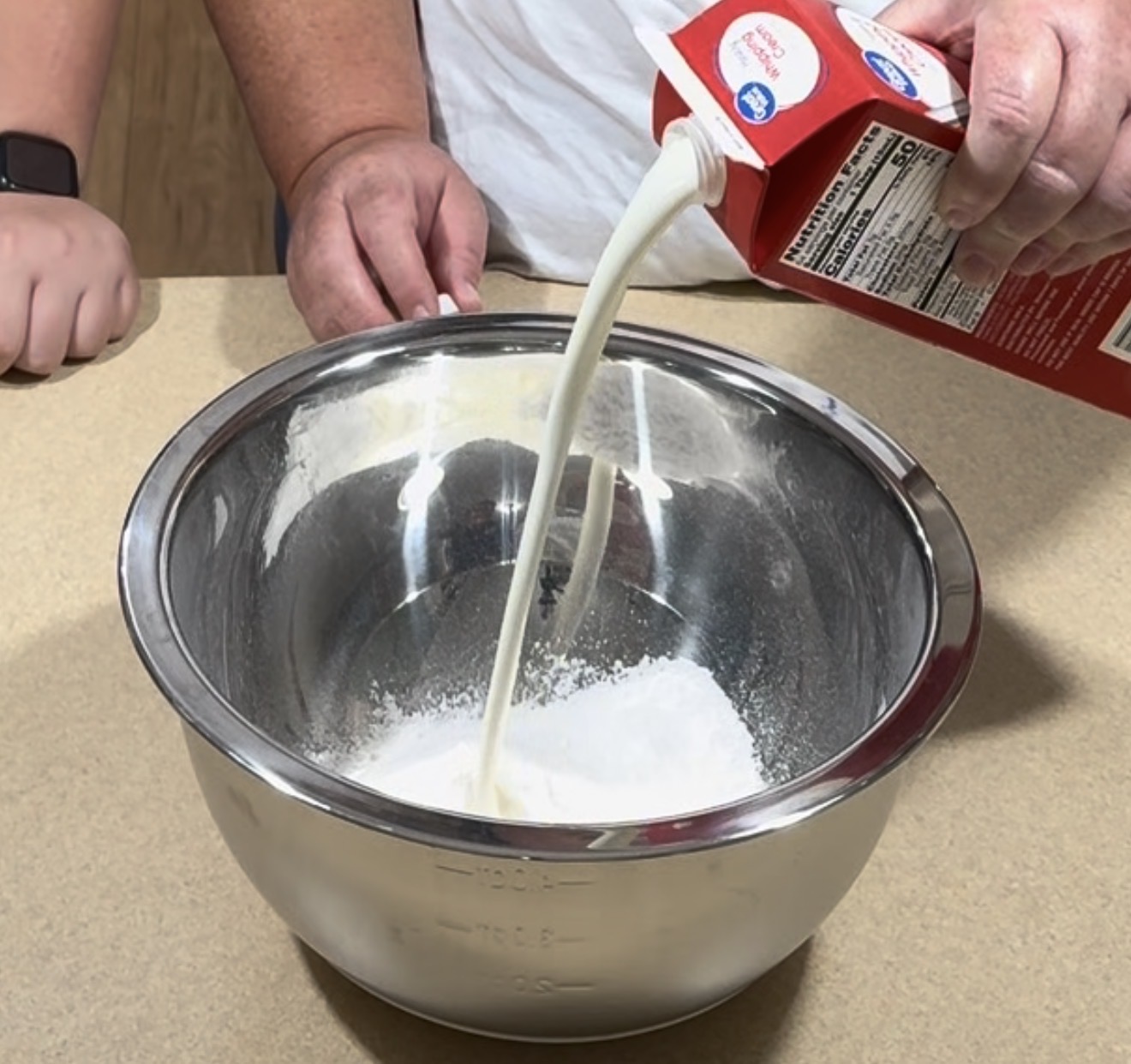 A person pours heavy whipping cream from a carton into a metal mixing bowl containing powdered sugar, preparing to make whipped cream. Another person stands nearby, partially visible.