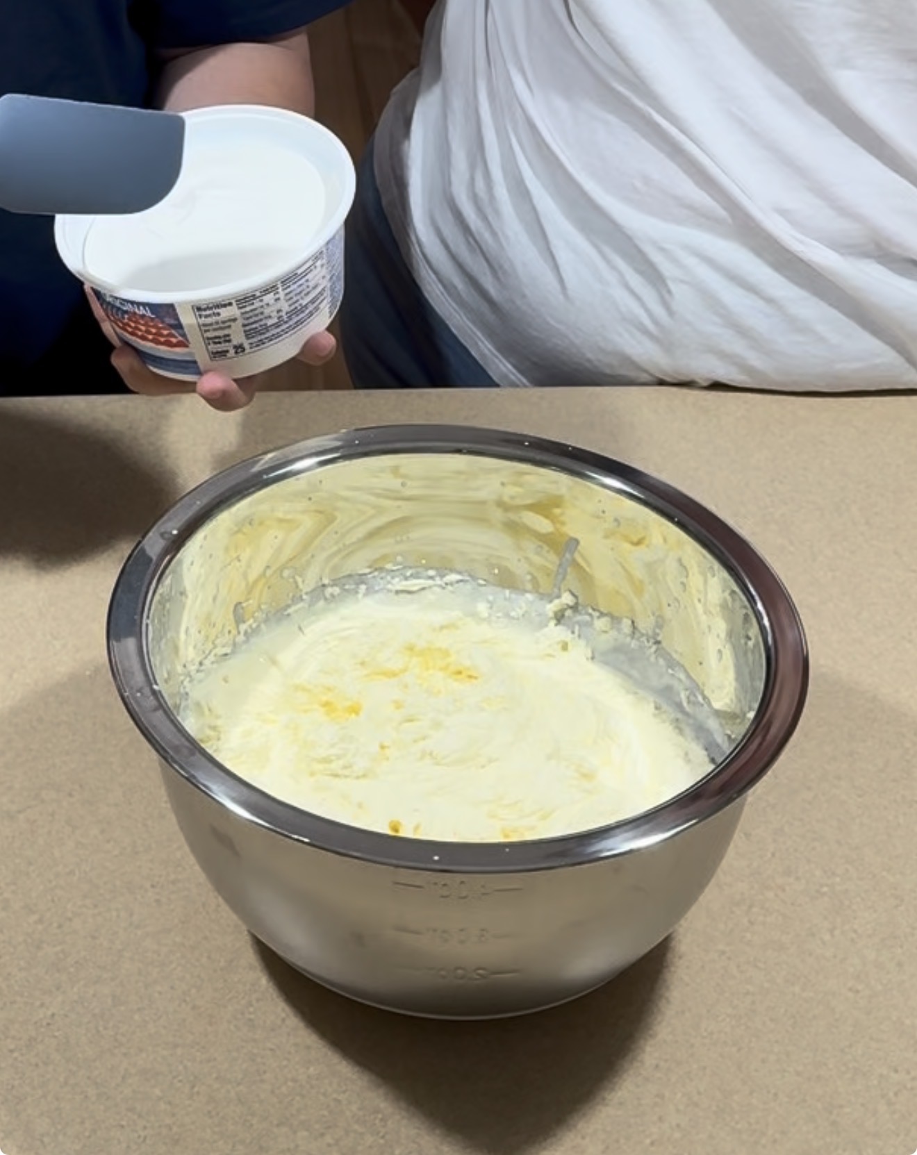 Two people stand by a counter; one holds a spatula and a container of sour cream, while a large metal bowl filled with creamy batter or mixture sits on the countertop.