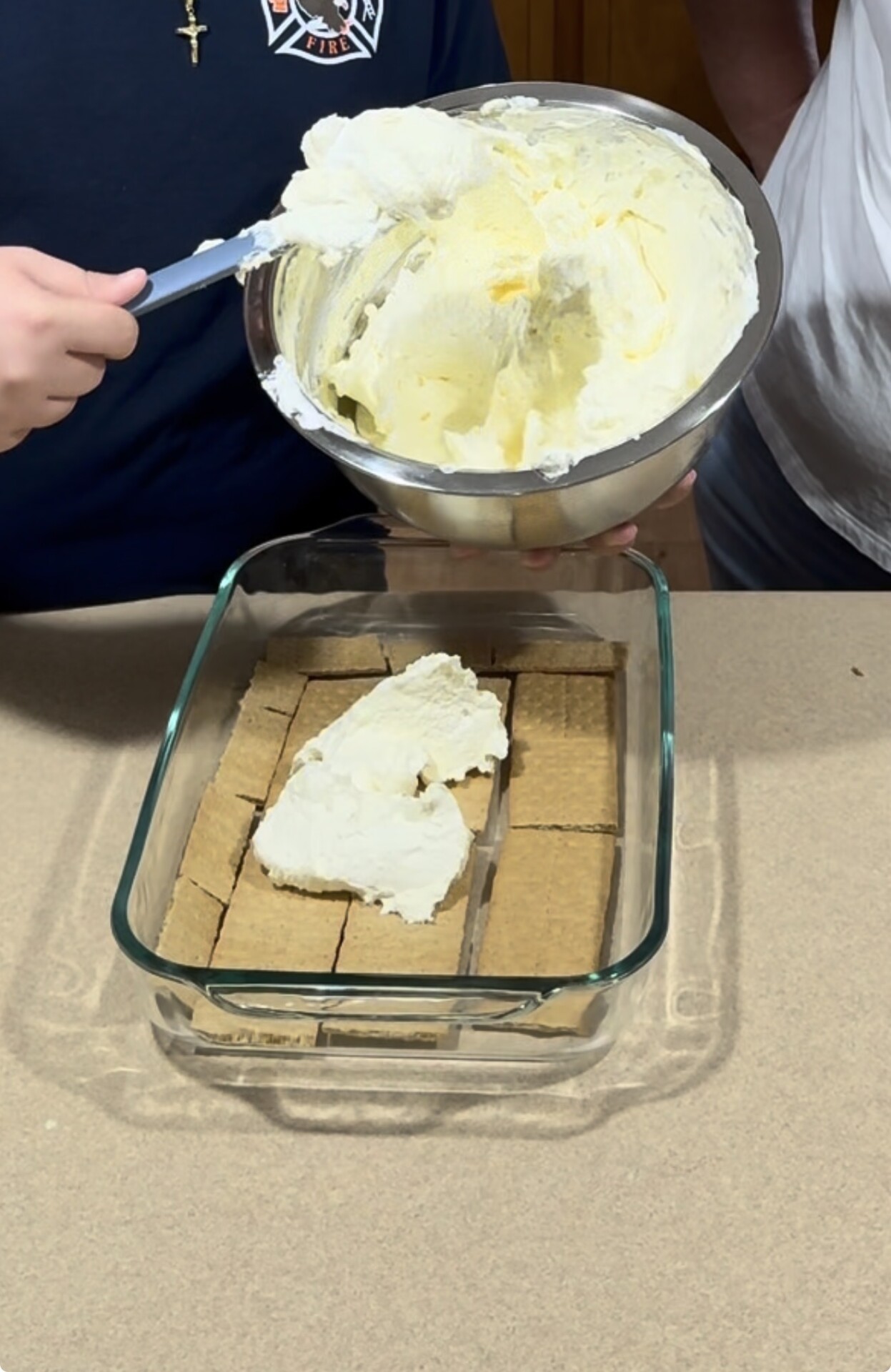 A person spreads creamy filling over a layer of graham crackers in a glass baking dish. Another person holds the bowl of filling above the dish. Both are working on a kitchen counter.