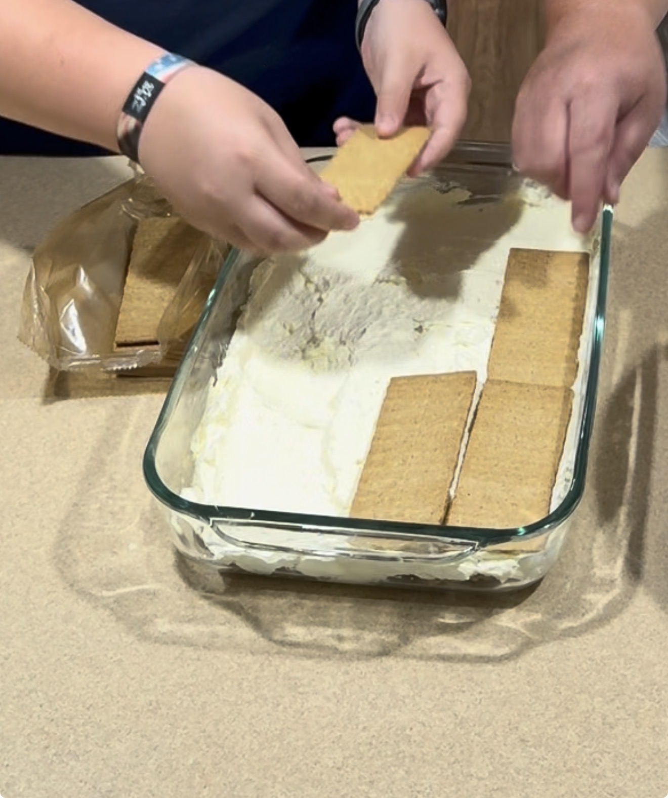 Two people place rectangular graham crackers into a glass baking dish filled with a creamy mixture, preparing a layered dessert on a kitchen counter.