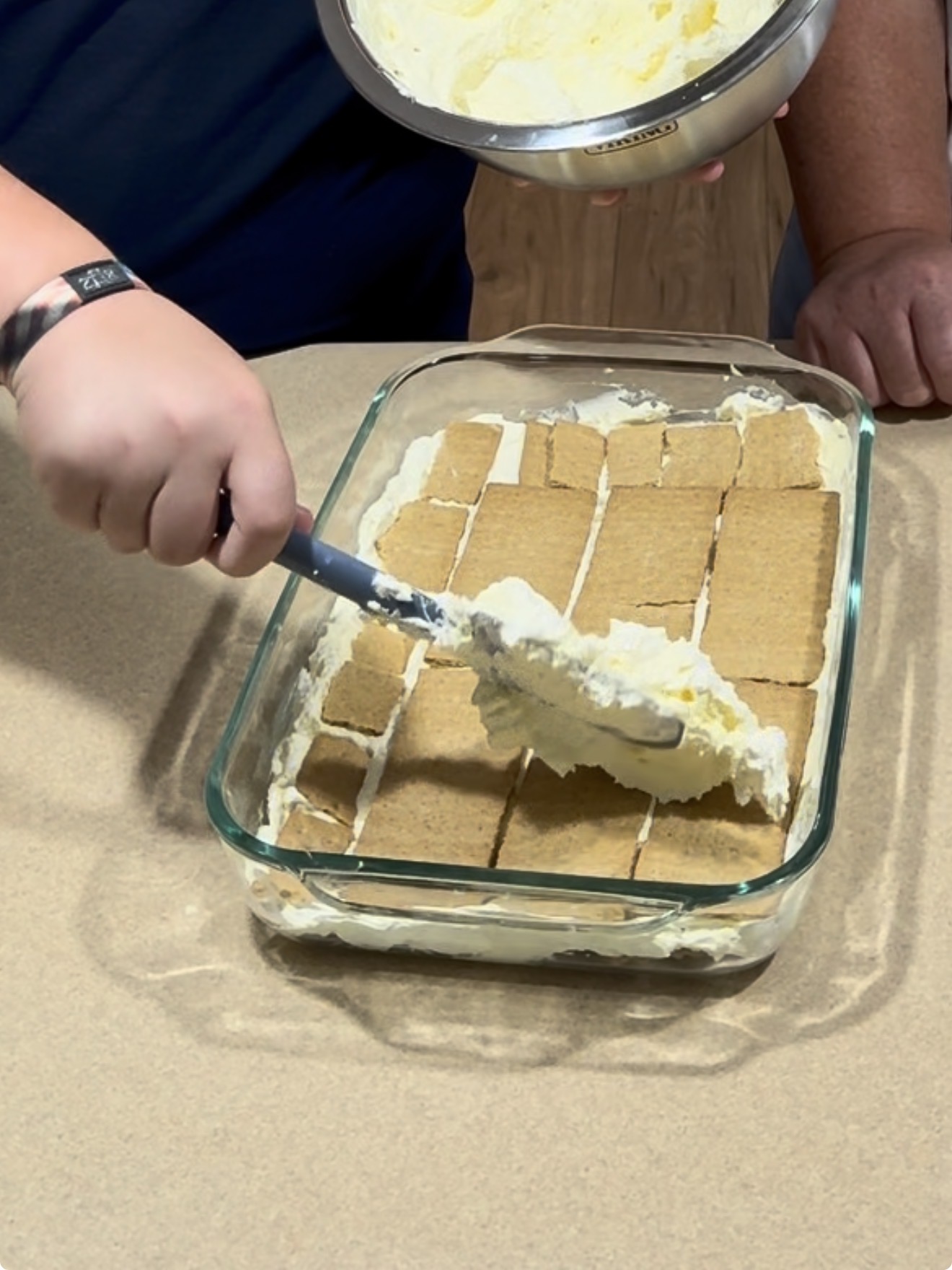 A person spreads creamy filling over a layer of rectangular graham crackers in a glass baking dish, preparing a dessert on a kitchen counter.