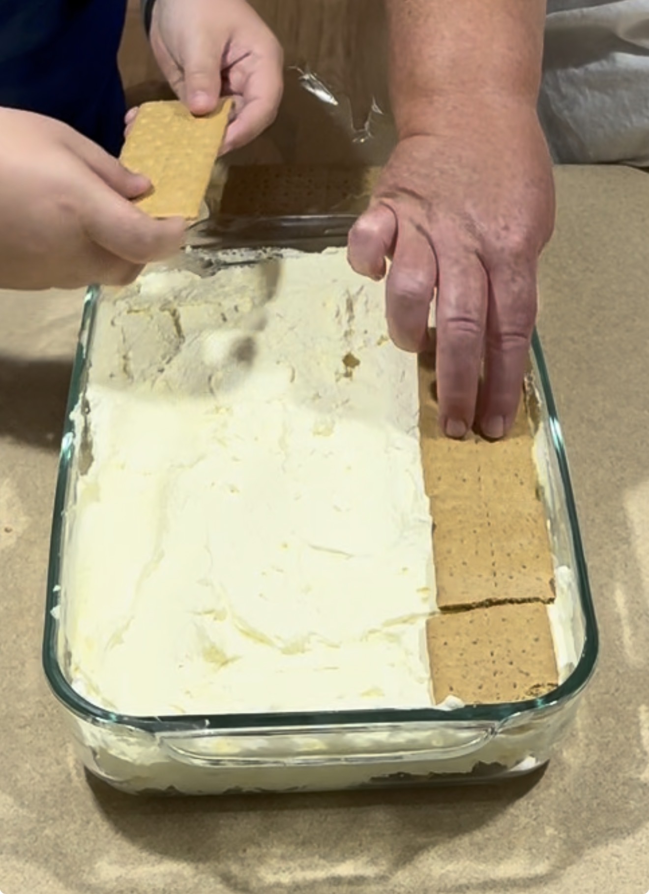 Two people are placing rectangular graham crackers on top of a layer of creamy filling in a glass baking dish, preparing a dessert on a brown surface.