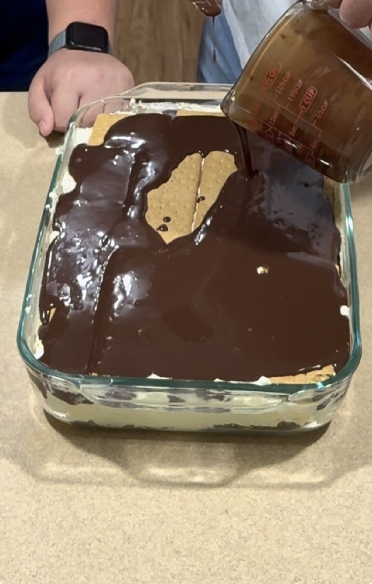 A hand pours chocolate topping over a layered dessert with graham crackers and cream in a glass baking dish, while another person’s arm rests nearby on the counter.