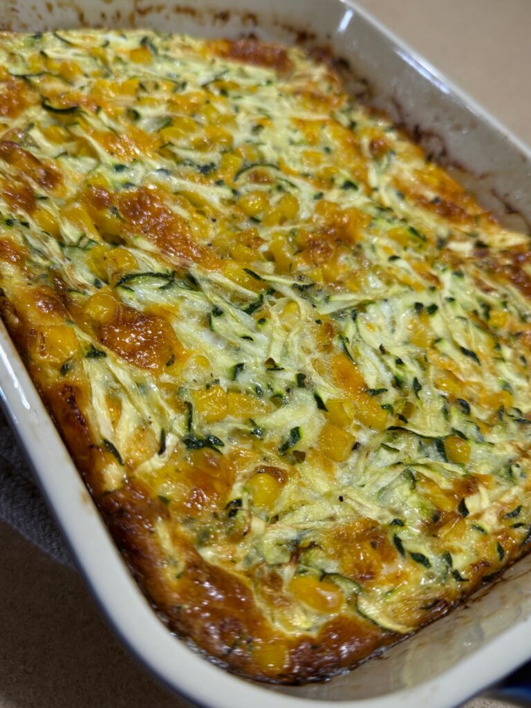 A close-up of a baked casserole in a white dish, showing a golden-brown top with visible pieces of zucchini and yellow corn mixed throughout.