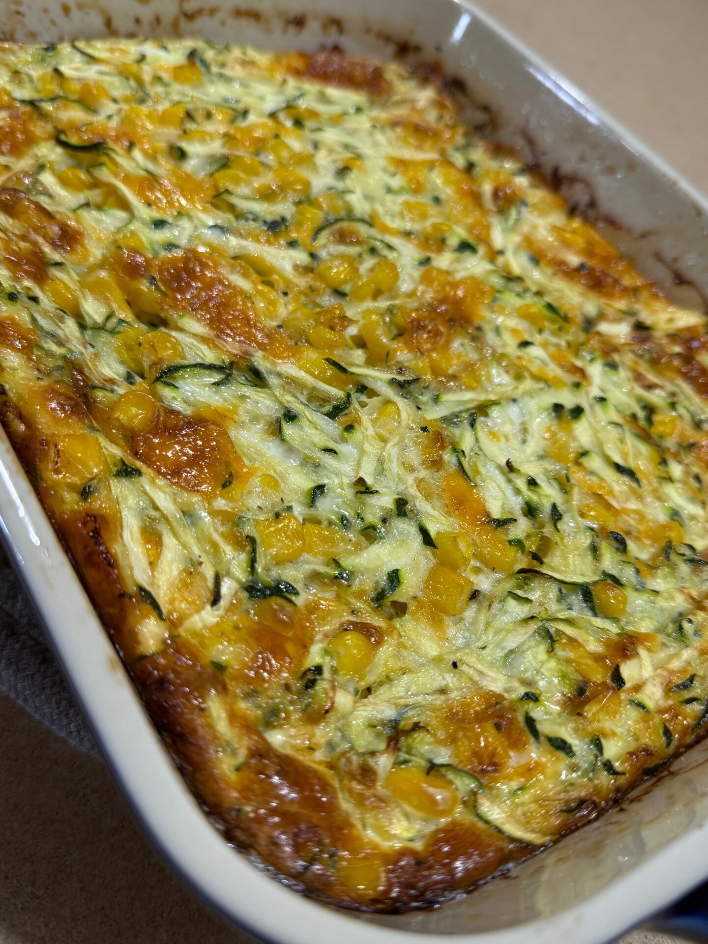 A close-up of a baked casserole in a white dish, showing a golden-brown top with visible pieces of zucchini and yellow corn mixed throughout.