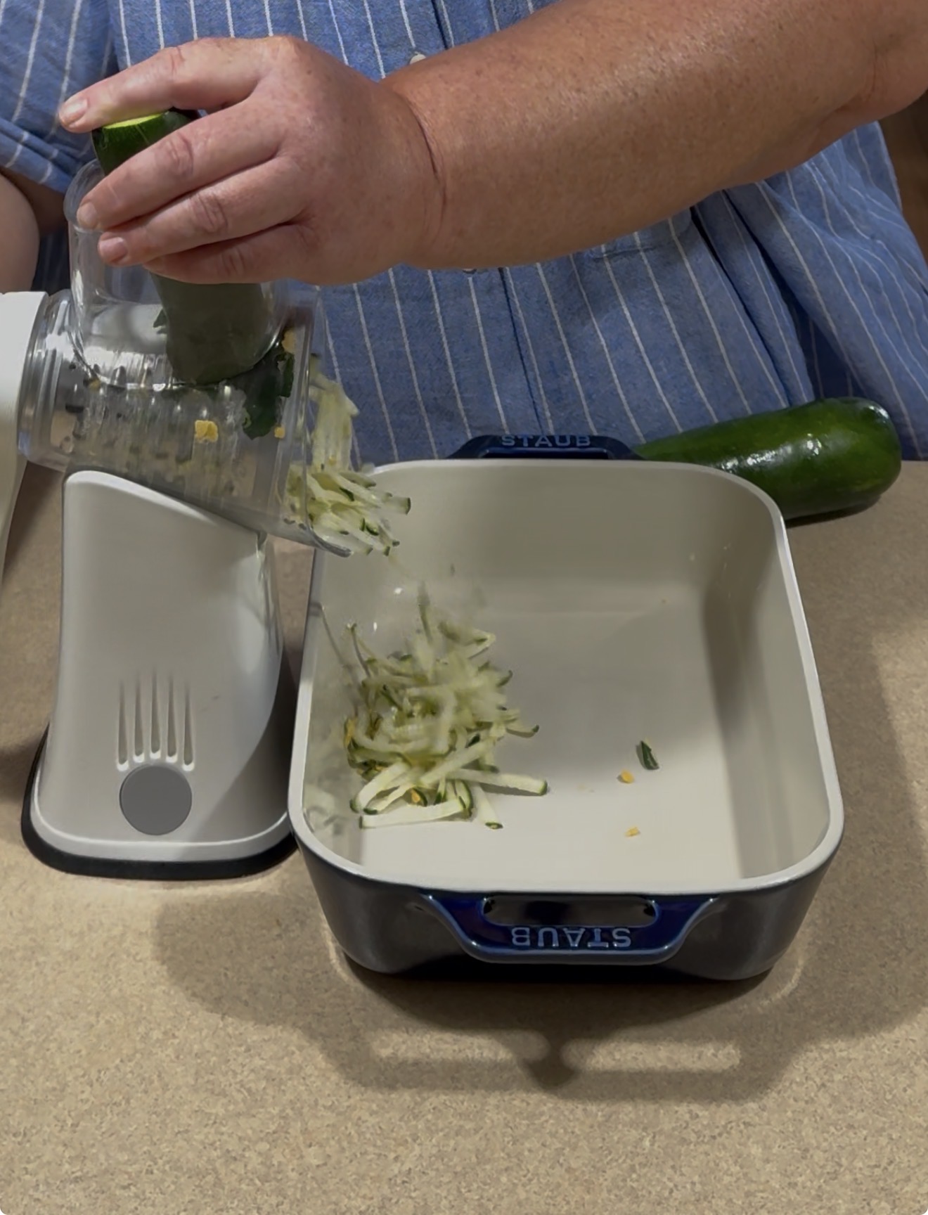 A person grates zucchini into a blue and white baking dish using a food processor. Another whole zucchini is on the counter beside the dish. The person is wearing a blue striped shirt.