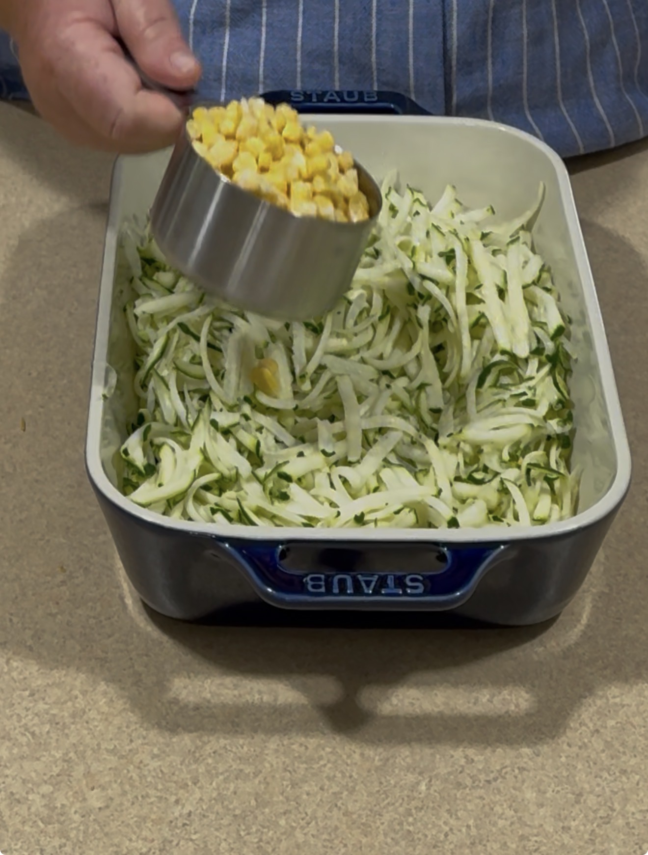 A person in a blue striped apron adds a cup of corn to a baking dish filled with shredded zucchini on a kitchen counter.