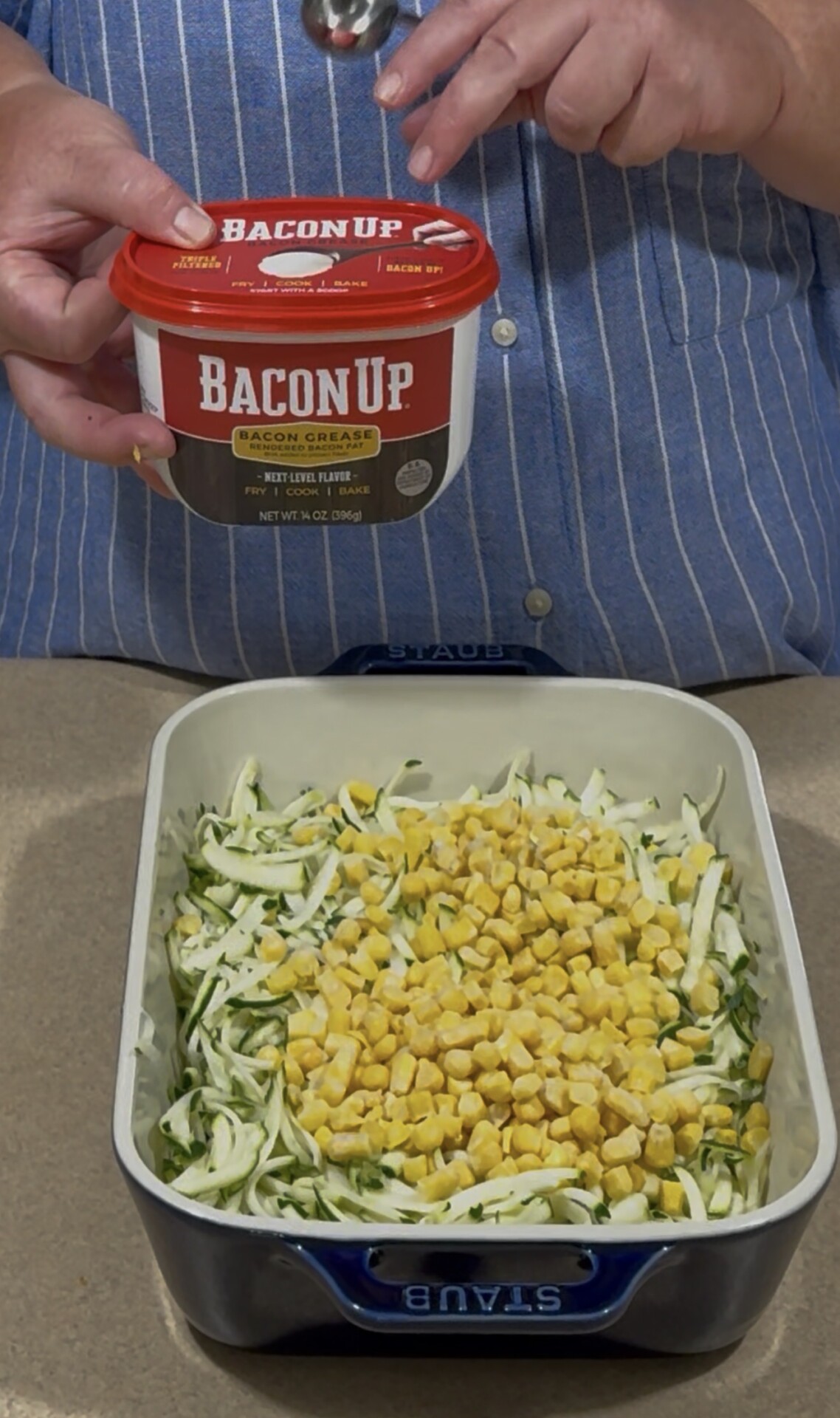 A person in a blue striped shirt holds a container of Bacon Up bacon grease above a baking dish filled with shredded cabbage and frozen corn.