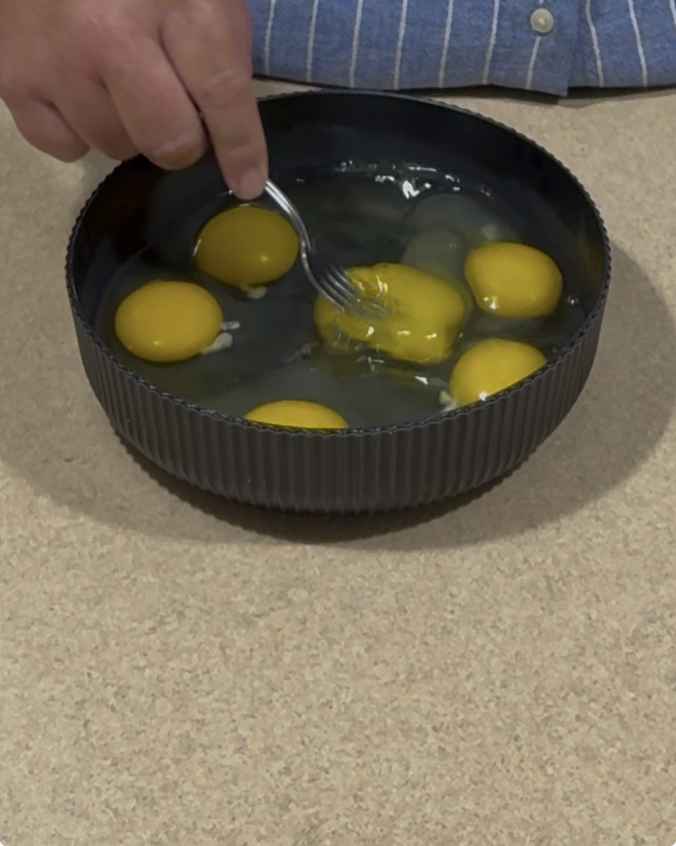 A person uses a fork to whisk several raw eggs in a black bowl on a beige countertop.