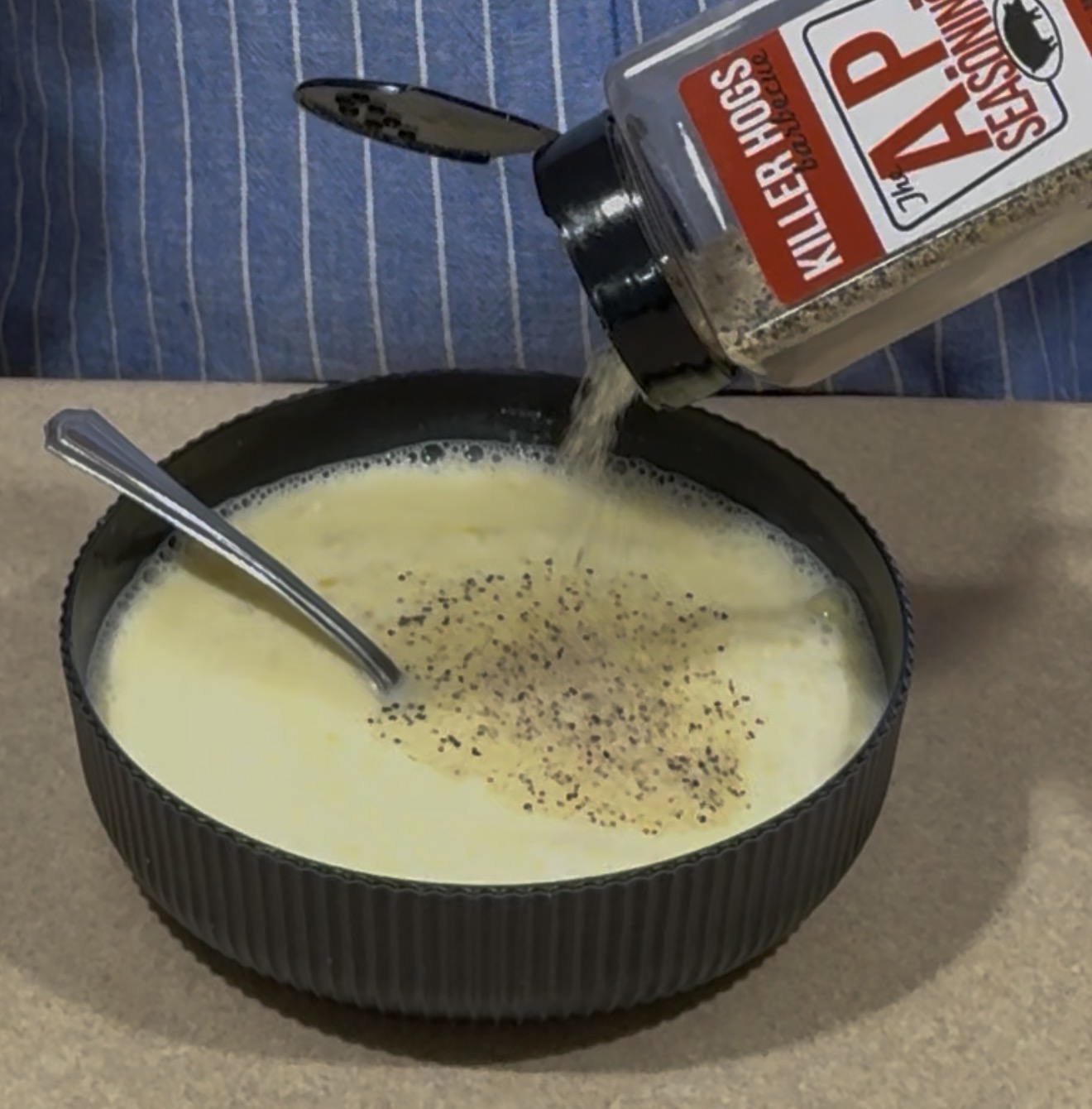 A person sprinkles seasoning from a Killer Hogs AP Seasoning bottle into a bowl of creamy soup, with a spoon resting inside the bowl.