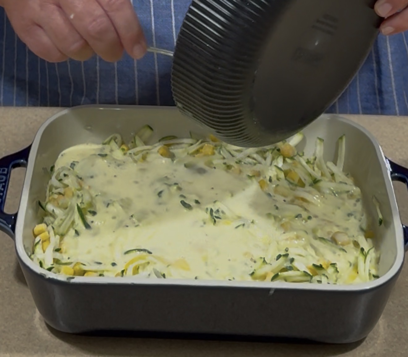 A person pours a creamy mixture from a black bowl over shredded vegetables in a blue rectangular baking dish on a kitchen counter.
