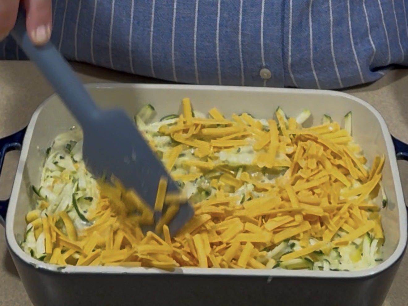 A person wearing a blue striped shirt uses a spatula to spread shredded cheddar cheese over a creamy zucchini casserole in a rectangular baking dish.