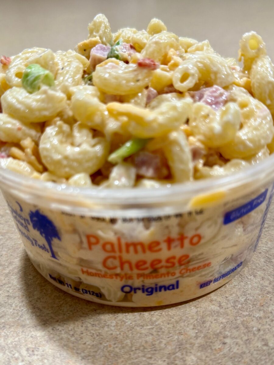 A close-up of macaroni salad with chopped vegetables in a container labeled Palmetto Cheese, Homestyle Pimento Cheese, Original. The pasta is creamy and the container sits on a beige surface.