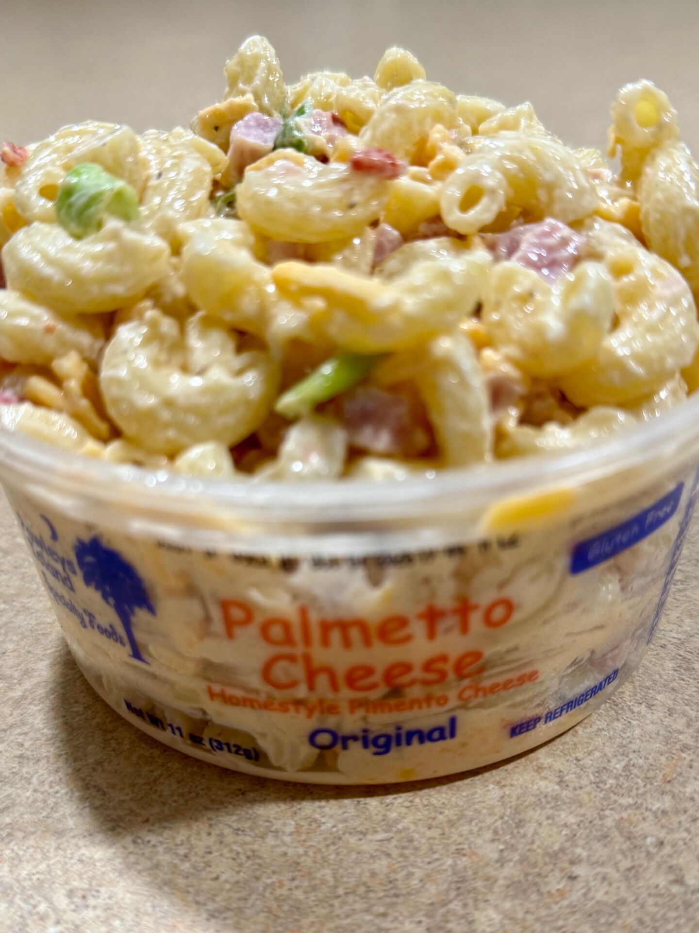 A close-up of macaroni salad with chopped vegetables in a container labeled Palmetto Cheese, Homestyle Pimento Cheese, Original. The pasta is creamy and the container sits on a beige surface.