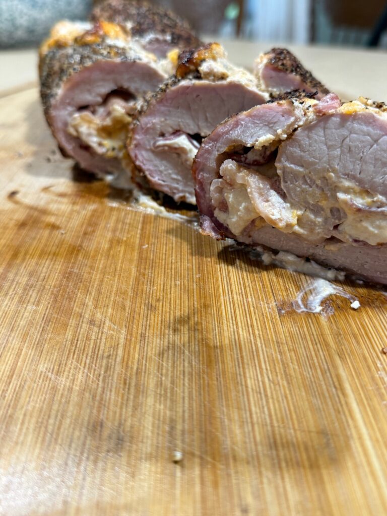 Sliced stuffed pork tenderloin on a wooden cutting board, showing seasoned meat with a creamy filling. The image focuses on the texture and detail of the meat and stuffing.