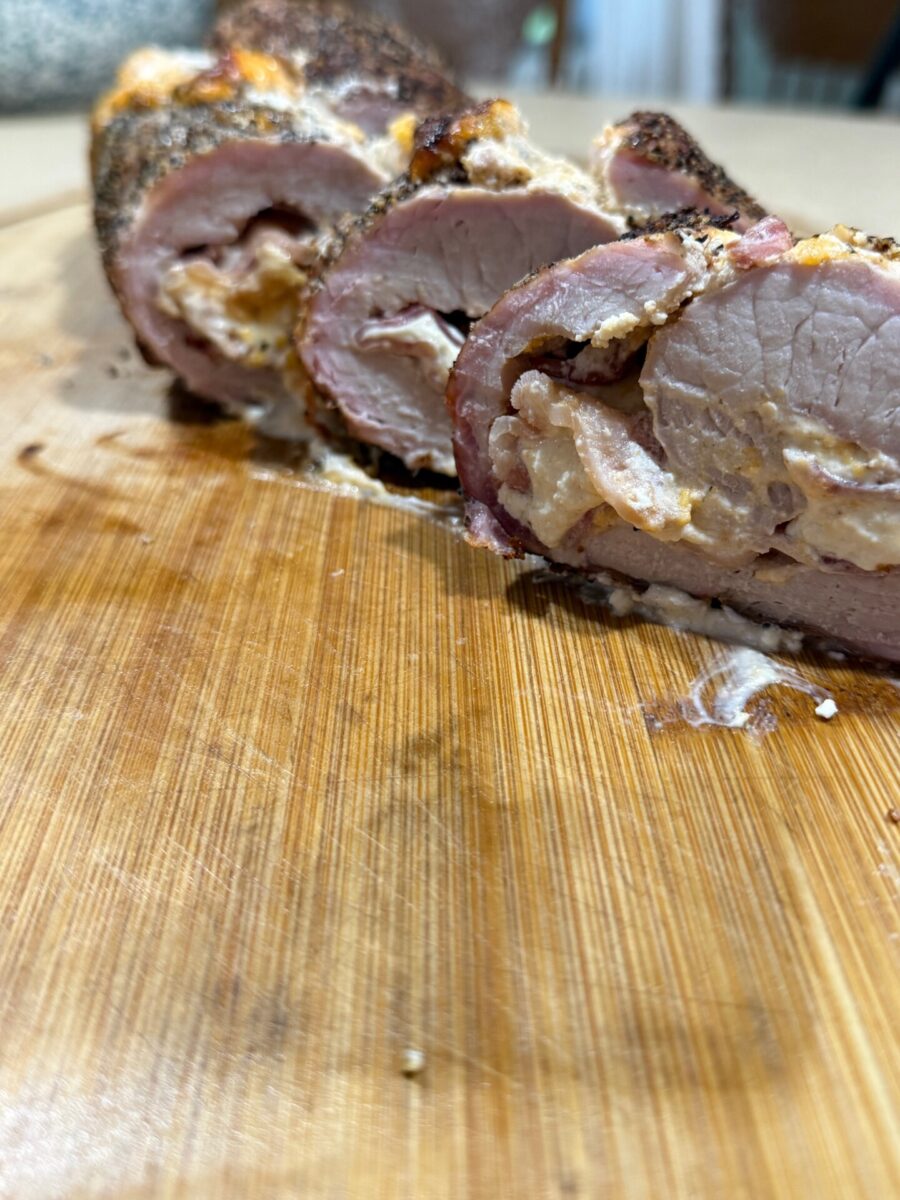 Sliced stuffed pork tenderloin on a wooden cutting board, showing seasoned meat with a creamy filling. The image focuses on the texture and detail of the meat and stuffing.