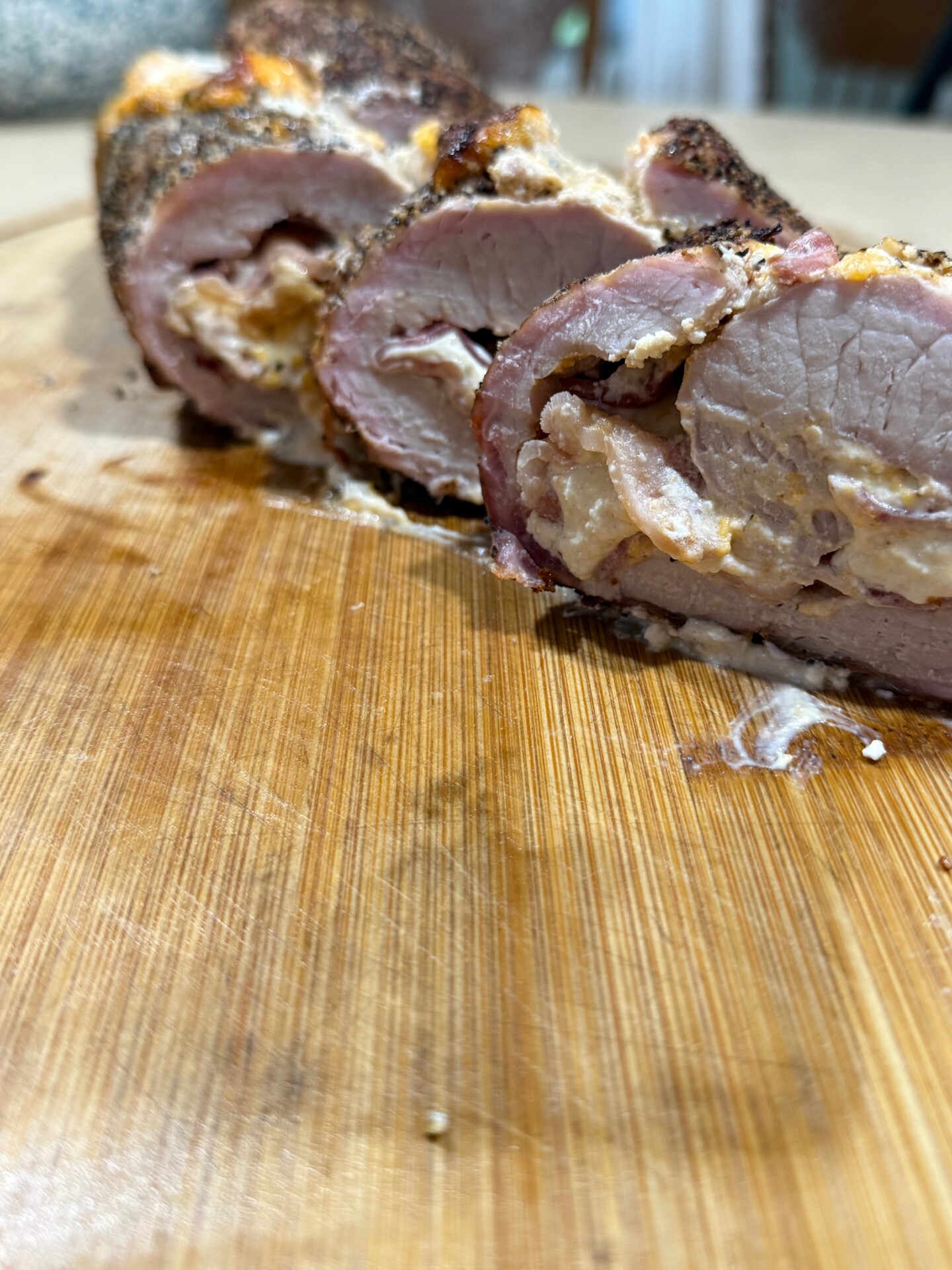 Sliced stuffed pork tenderloin on a wooden cutting board, showing seasoned meat with a creamy filling. The image focuses on the texture and detail of the meat and stuffing.