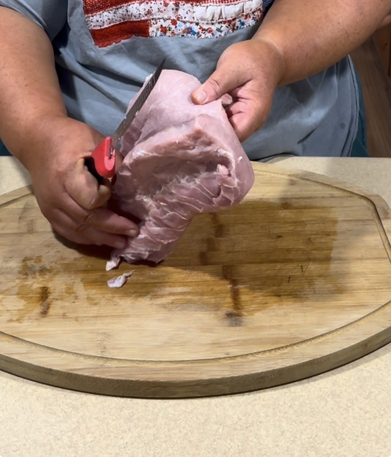 A person holding a large piece of raw pork on a wooden cutting board, preparing to slice it with a knife. The person is wearing a grey T-shirt.