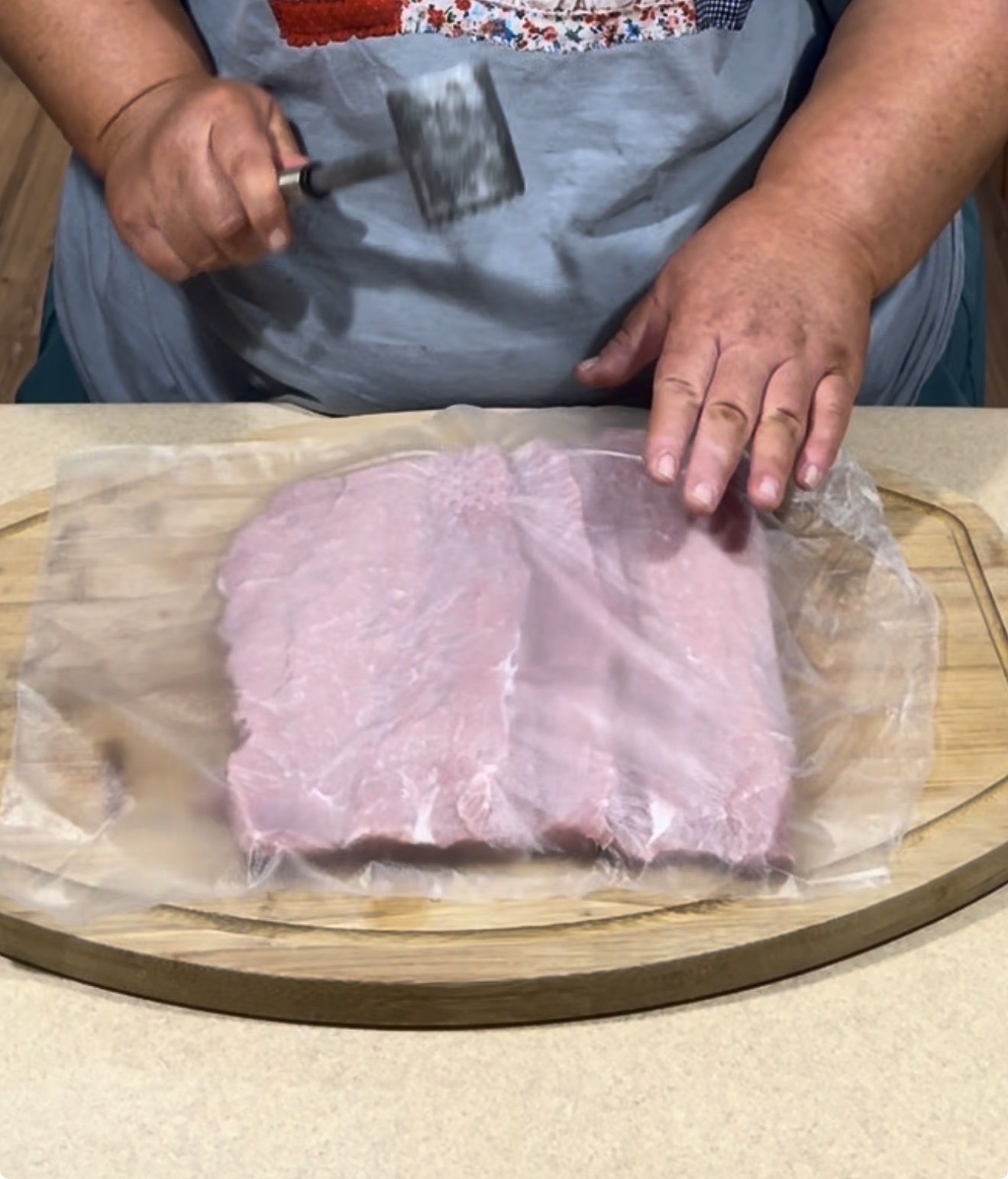 A person uses a meat tenderizer to pound a piece of raw meat covered with plastic wrap on a wooden cutting board.