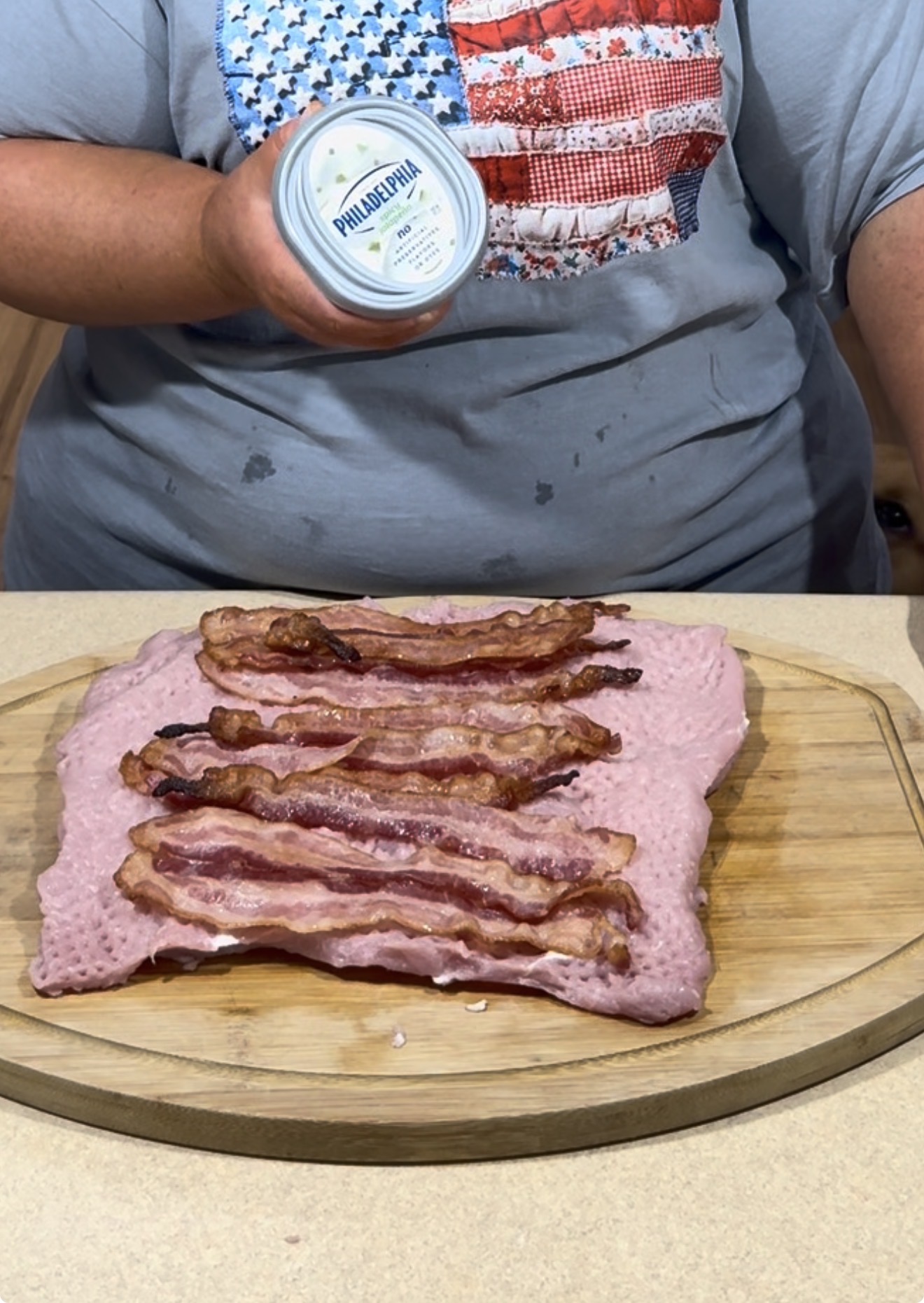 A person in a gray shirt with an American flag design holds a tub of Philadelphia cream cheese over a cutting board with a pink meat slab topped with strips of cooked bacon.