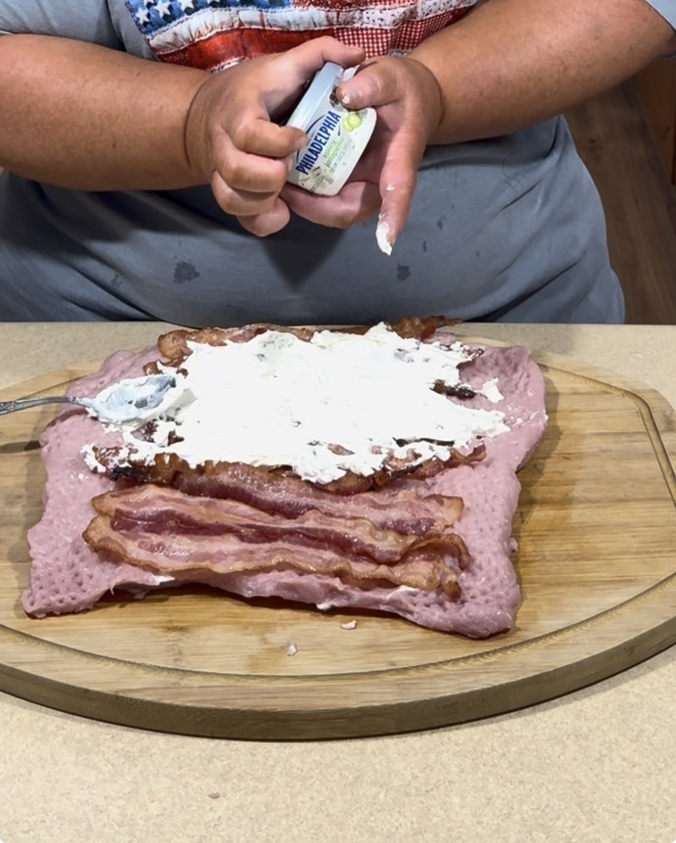 A person spreads cream cheese onto a layer of cooked bacon and processed meat laid out on a wooden cutting board. A spoon rests on top of the cream cheese.